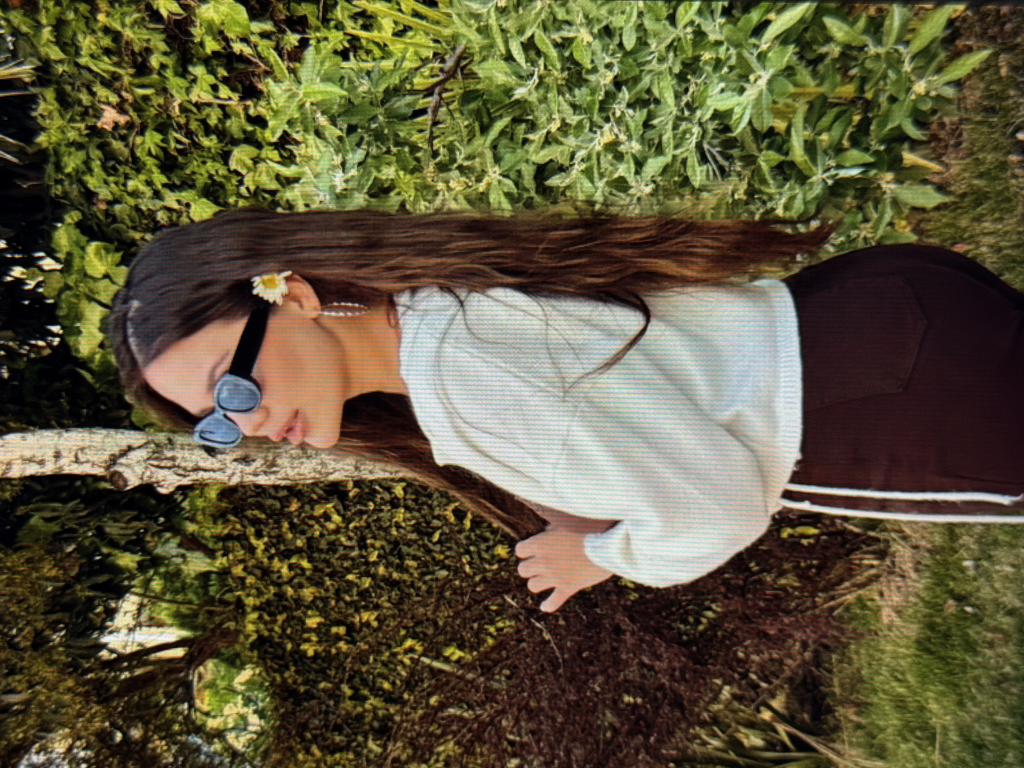 A young woman with long brown hair stands outdoors, turned to her left, in front of a large tree and a dense green hedge. She wears black sunglasses, a white cropped hoodie with white drawstrings, and dark maroon pants. A small white flower is tucked behind her right ear, and she has a silver hoop earring in her visible ear. Her right hand is held up near her head, near her hair. The background includes green grass and more foliage. The scene is set in a park or garden in the city of Sirius, Russia.