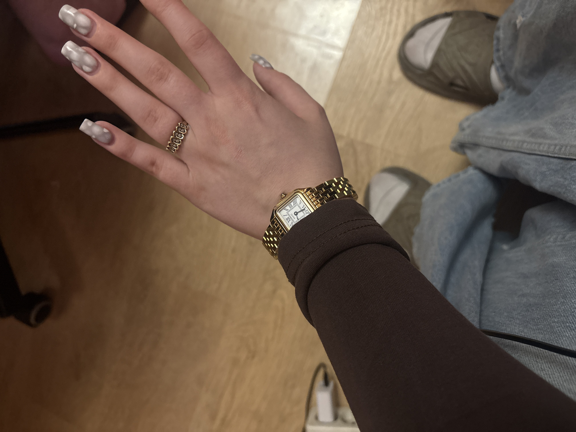The image shows a close-up of a person's left arm and hand, wearing a brown long-sleeved shirt and blue jeans. On the wrist, there is a gold watch with a white face and Roman numerals. The person is wearing a gold ring with multiple circular elements on their right ring finger. The nails are painted with a marbled gray and white design. The background consists of a wooden floor. There is no indication of the city of Khimki, Russia, or any other specific location.