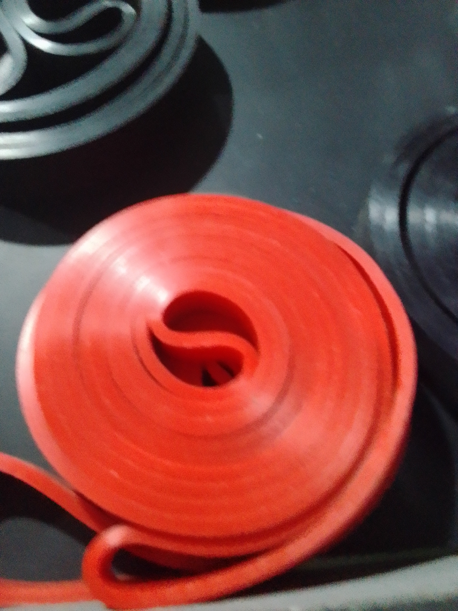 This is a close-up, slightly blurry, eye-level shot of a roll of bright red, rubbery material, possibly a gasket or seal, lying on a dark grey surface. The roll is coiled tightly, forming a circular shape with a prominent hollow center. The material appears smooth and slightly reflective, with some subtle creases and variations in its surface texture. The edges of the roll are rounded and appear somewhat flexible.
In the background, to the left, part of another object is visible. It is dark grey or black and has a layered, spiral-like design. To the right and further back, another black object with a curved edge is partially visible. The lighting is dim, casting soft shadows and highlighting the curves of the red roll. There is no visible text in the image. The scene appears to be indoors, possibly a workshop or storage area. There are no people or any indications of the time of day or weather. The overall impression is of raw materials or components.
