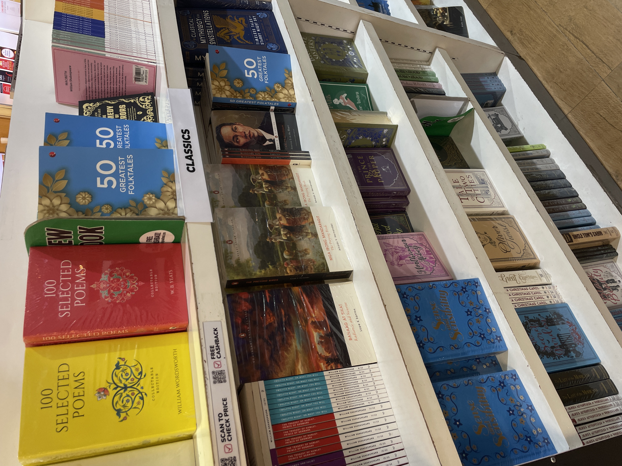The image shows a shelf of books in a bookstore, with several titles prominently displayed. On the top shelf, a yellow book titled "100 Selected Poems" by William Wordsworth is visible, next to a red book with the same title but by W.B. Yeats. To the right, blue books with "50 Greatest Folktales" are displayed. Below this, a row of books from the "William Shakespeare" collection is lined up. Further down, "Sense and Sensibility" is featured in blue covers, and a book titled "The Secret Garden" is also visible. In the lower right section of the image, a shelf displays books including "Oliver Twist" and "Tale of Two Cities." The overall environment suggests a retail setting, likely a bookstore. The presence of a "SCAN TO CHECK PRICE" sign with a QR code indicates a modern retail approach. The flooring visible at the bottom of the image appears to be wooden planks. Without additional context, it is not possible to pinpoint the exact location within Davao City, Philippines, but the types of books displayed suggest it's a general bookstore or a specific section dedicated to classics and literature.