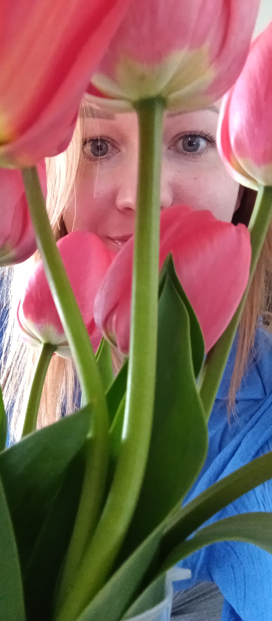 A woman with blonde hair and blue eyes is partially visible behind a bouquet of pink and green tulips. She is wearing a blue shirt. The location is unknown.