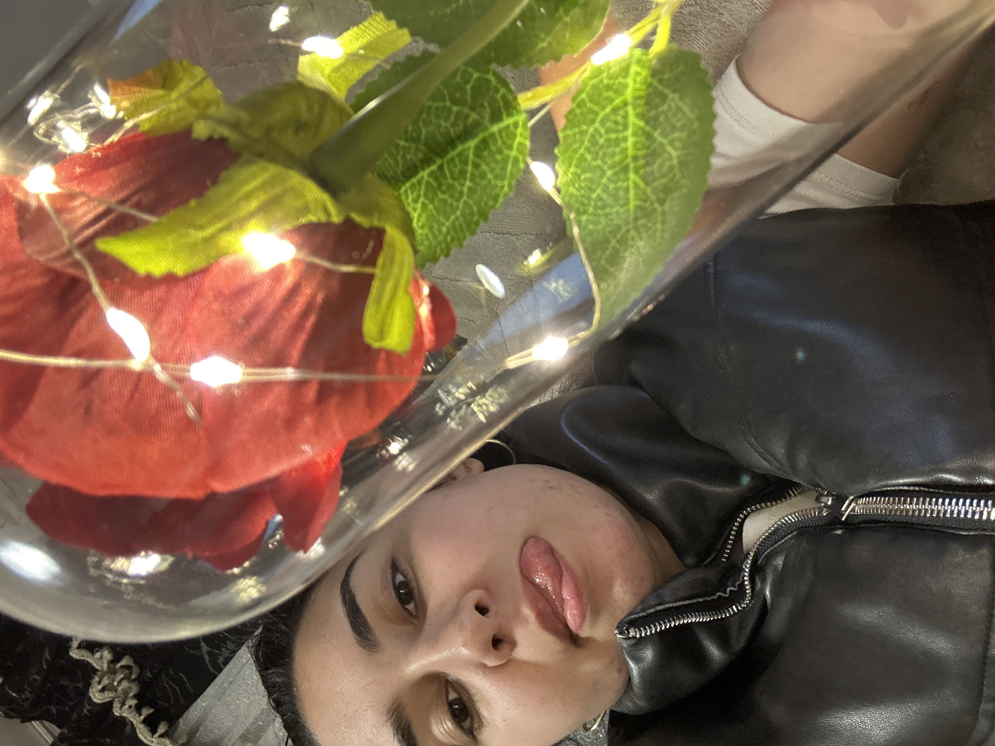 A woman lies on a surface, possibly a bed or the floor, wearing a black leather jacket with a partially zipped zipper. She is holding a glass dome containing a red rose with green leaves and string lights. The woman is looking at the camera with her tongue sticking out. The location in the city of Burgas, Bulgaria cannot be determined from the image.