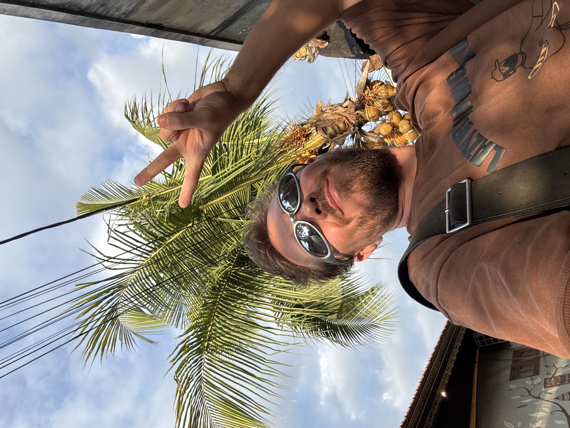 A low-angle shot captures a man in North Kuta, Indonesia, posing against a backdrop of a palm tree and a partly cloudy sky. He wears sunglasses and a brown shirt and holds up a "peace" hand gesture. Behind the man and the palm tree, a building is visible, with the fronds of the palm tree nearly obscuring the building. Power lines run above the palm tree. The scene conveys a tropical, casual vibe.