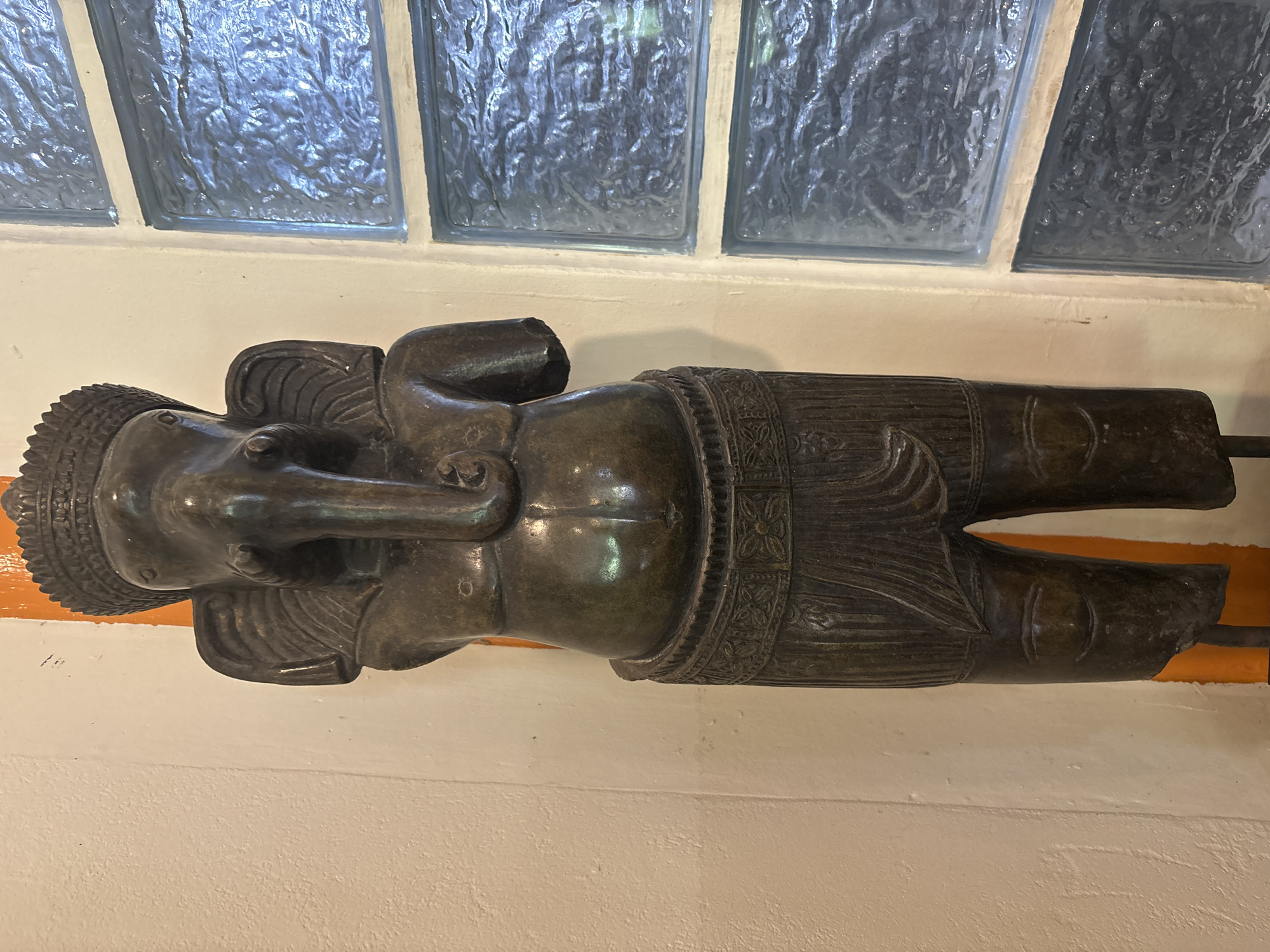 The image shows a full-body, dark bronze statue of Ganesha, the Hindu deity with an elephant head. The statue is standing and is positioned in front of a cream-colored wall and to the right of a bright orange vertical support. To the right of the statue, there is a wall composed of blue glass blocks, creating a textured and translucent background. The statue itself is intricately detailed, with a crown on its head, large elephant ears, and a prominent trunk. It has a stout torso and is wearing a decorative band around its waist, from which hangs a draped lower garment. The legs are depicted as somewhat stylized, with etched lines suggesting knees and feet. The statue is lit from the front, casting subtle shadows and highlighting the metallic sheen of the bronze. The overall environment appears to be indoors, possibly a temple, museum, or a private collection, given the display context. The location is specified as Chalong, Thailand.