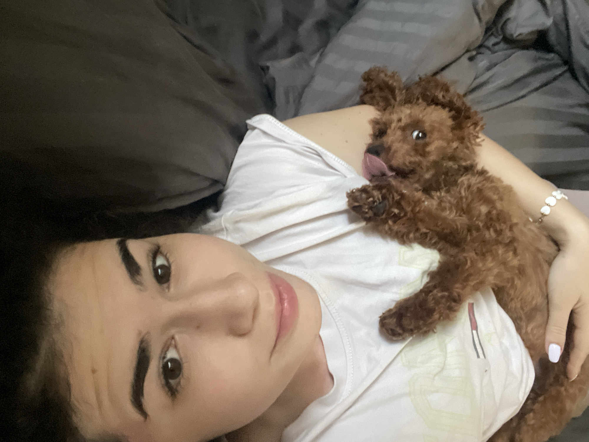 A woman and a dog are lying in bed. The woman is on her back, and the dog is lying on her chest. The woman is wearing a white shirt. The dog is brown and has a long tongue. The bed has gray sheets and a gray blanket. There is no indication that the image was taken in Sofia, Bulgaria.
