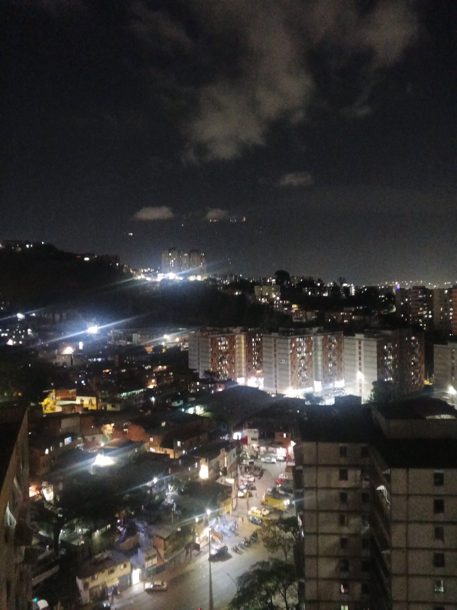 The image depicts a nighttime urban landscape in Caracas, Venezuela. The foreground features a densely populated area with low-rise buildings, interspersed with illuminated streets and vehicular traffic. A multi-story apartment building is visible on the right side of the image. In the middle ground, a series of high-rise residential buildings with lit windows creates a distinct skyline. The background displays a hill with scattered lights, topped by additional buildings. The sky is dark with cloud cover. Overall, the scene presents a typical urban setting at night.