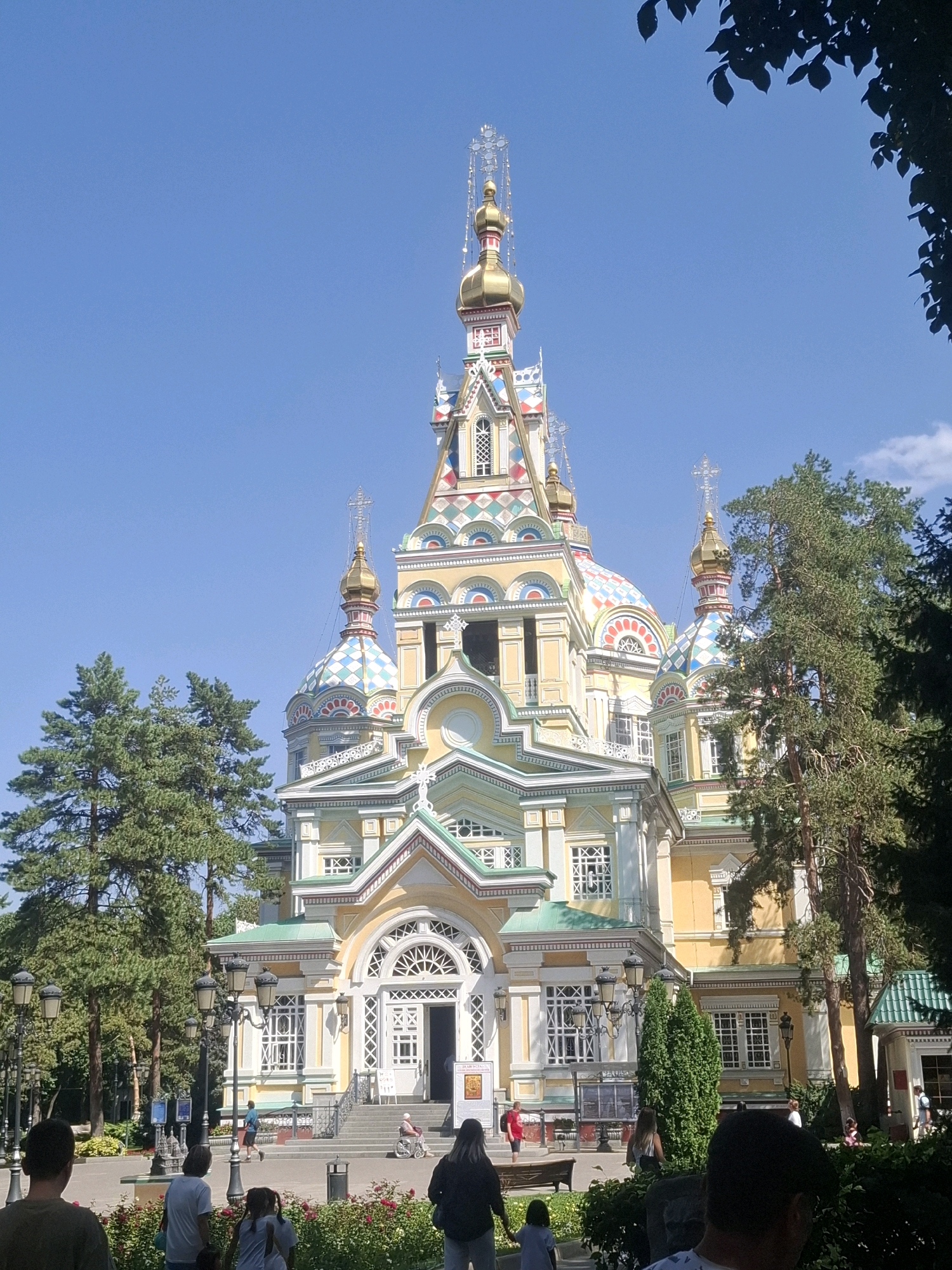The image shows Zenkov Cathedral, a wooden Orthodox church in Panfilov Park, Almaty, Kazakhstan. The cathedral, painted in bright yellow and white with a green roof, is intricately decorated with colorful geometric patterns on its domes and facades. Several people are visible in the foreground and on the steps of the cathedral. In the foreground, a group of people are standing, some facing away from the camera, and others are walking. One person is in a wheelchair being pushed up the steps. Tall trees, including pine trees, surround the cathedral. The sky is clear and blue. Street lamps are also present in the park area. The overall scene depicts a sunny day at a popular landmark.