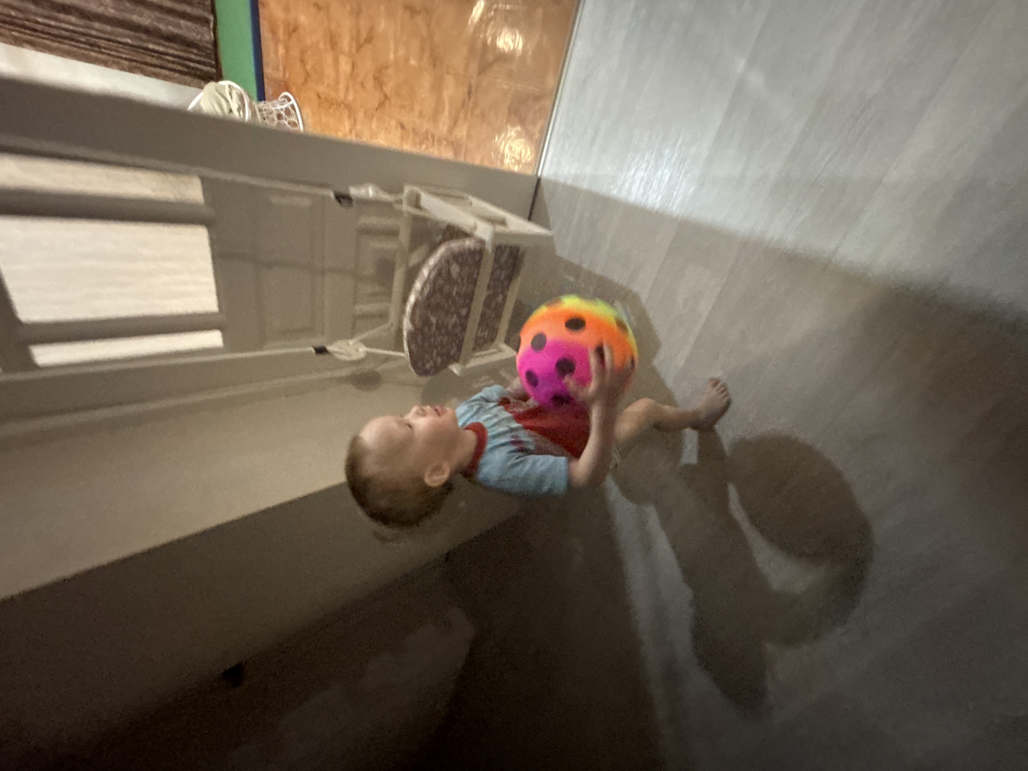A photograph depicts a young child lying on a light-colored floor, holding a brightly colored ball. The child is wearing a blue and red patterned shirt. The floor appears to be made of wood. A white structure, possibly a shelf or cabinet, is visible in the upper left. The background includes a wall with a wood-like pattern. The environment appears to be indoors, potentially a home. The location in the city of Ferma, Russia, is unknown.