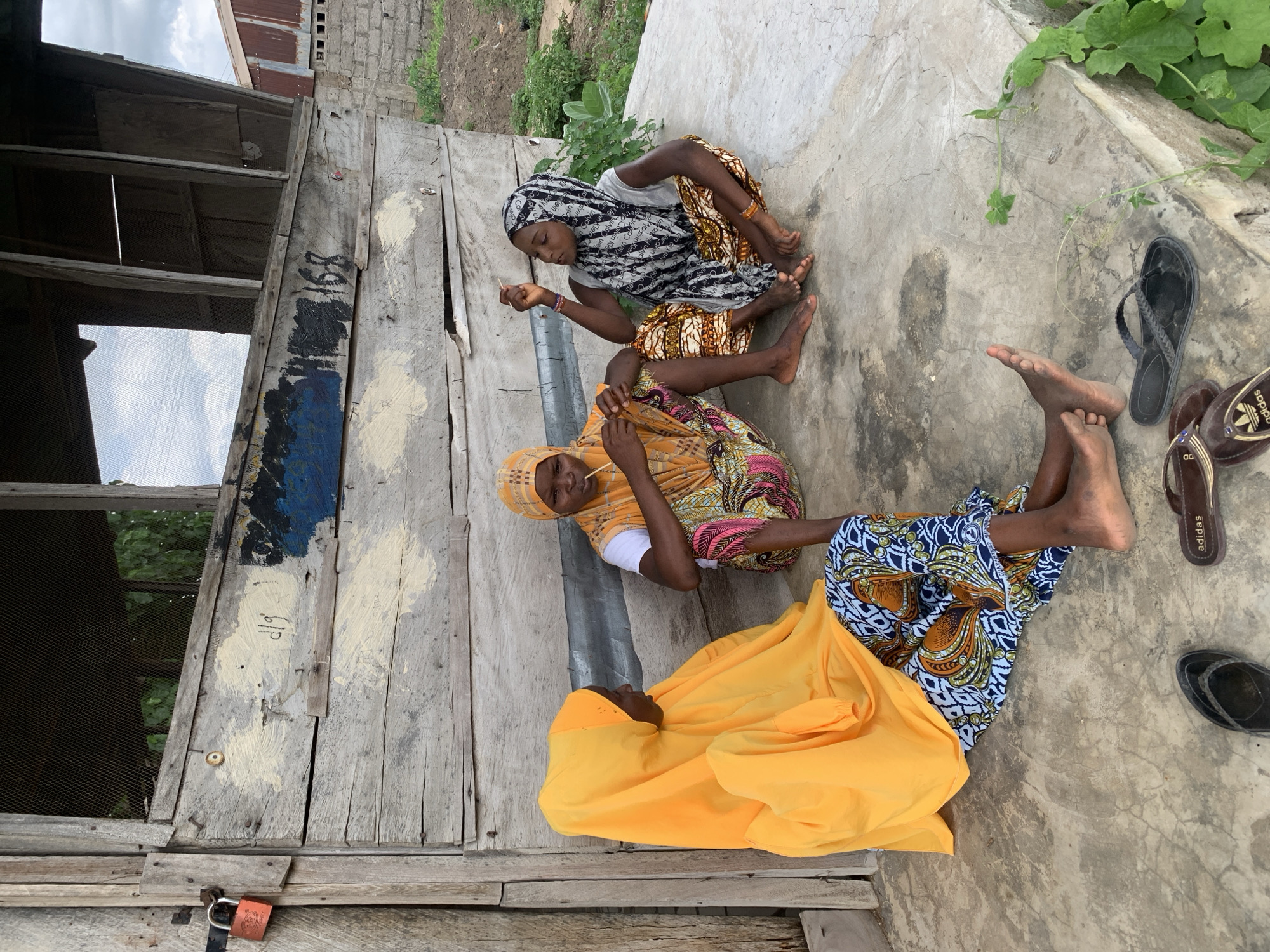 Three women are sitting on a concrete surface next to a weathered wooden structure in Adankolo, Nigeria. The women are wearing colorful patterned clothing and head coverings. Two of the women are holding small sticks. A pair of sandals is visible next to one of the women. The wooden structure has a window covered with wire mesh and a padlock.