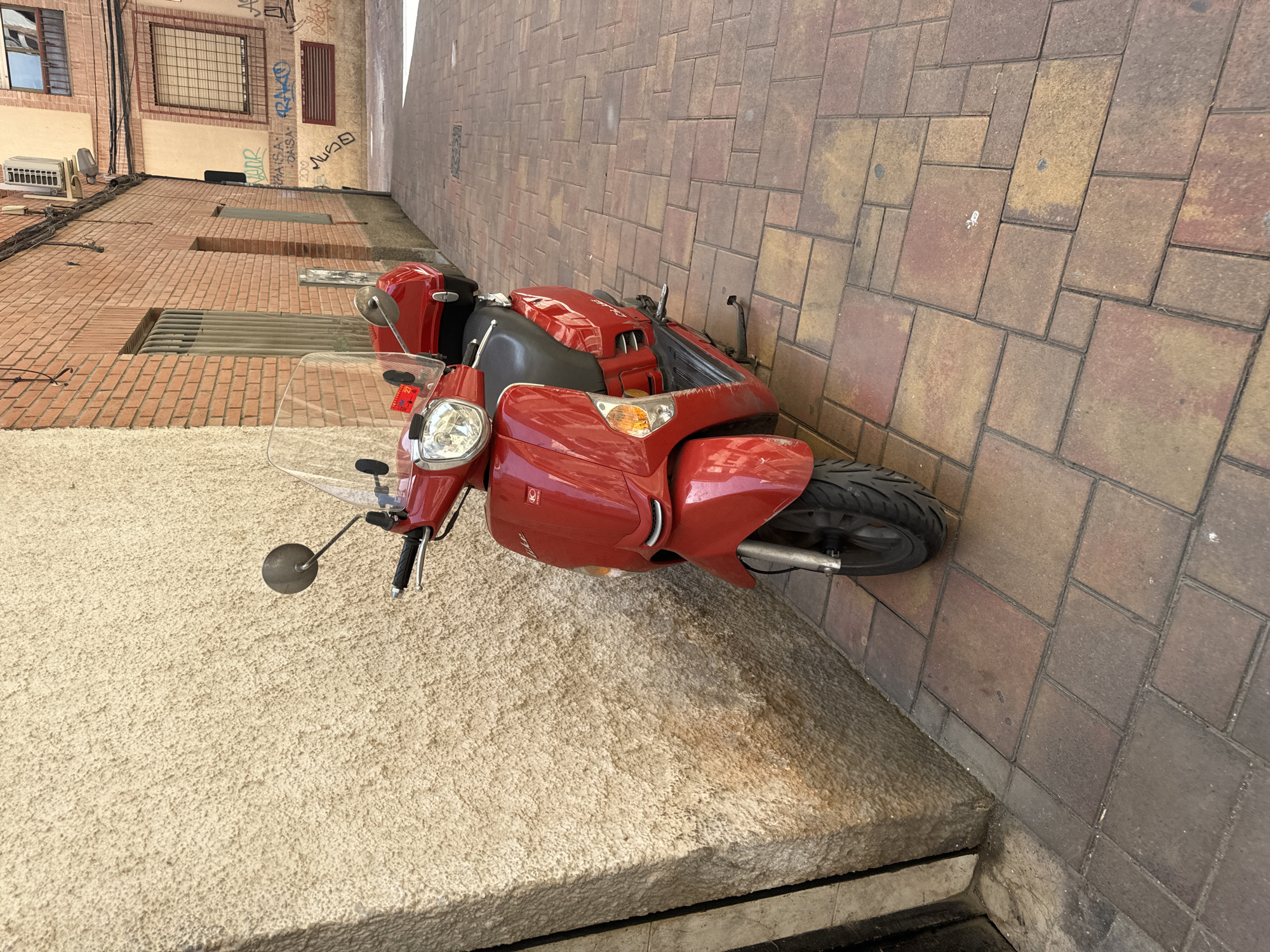 A red Kymco scooter is parked on a pedestrian walkway made of reddish-brown interlocking pavers. The scooter is positioned in front of a textured, light-colored stucco wall on the left side of the image. To the right of the scooter, a narrow alleyway opens up, lined with brick buildings. The buildings feature windows with metal grates and graffiti. An air conditioning unit is visible on one of the building facades, along with some wiring. The scene appears to be in a city, possibly in Murcia, Spain, given the architectural style and the presence of graffiti. The lighting suggests it is daytime, with sunlight casting shadows on the ground. There are no people visible in the image.