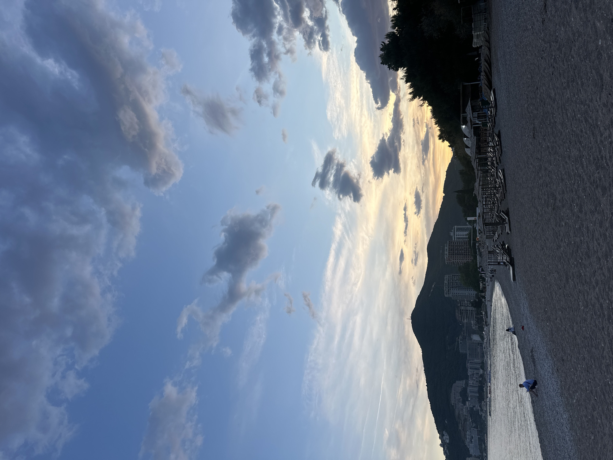 The image captures a beach scene in Budva, Montenegro, likely taken during early evening based on the sky's light. To the left, the sky shows a mix of blue and pale yellow with wispy clouds and dark patches. A tree line and rows of empty beach umbrellas are visible along the shoreline. In the background, a mountain rises, with urban buildings nestled at its base. Two individuals appear on the beach, one closer and one further, both near the water's edge. The beach has dark sand.