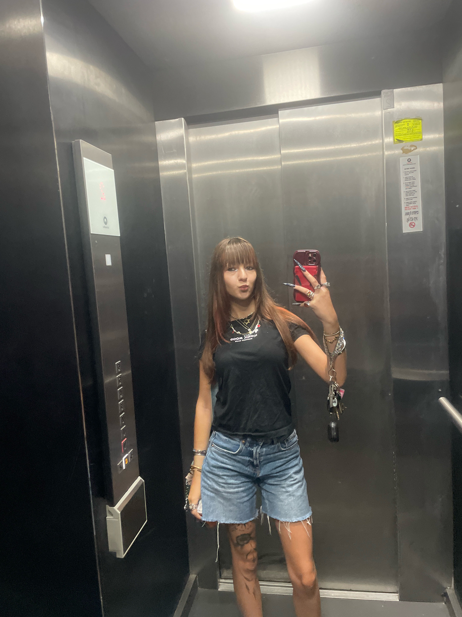 A young woman with long brown hair and bangs, wearing a black t-shirt, denim shorts, and jewelry, takes a mirror selfie inside an elevator. The elevator interior is stainless steel, with a control panel on the left displaying floor numbers and buttons. A yellow sticker is visible on the right-hand wall. The woman is holding a red smartphone in her right hand, and a set of keys hangs from her wrist. She appears to be in Raanana, Israel.
