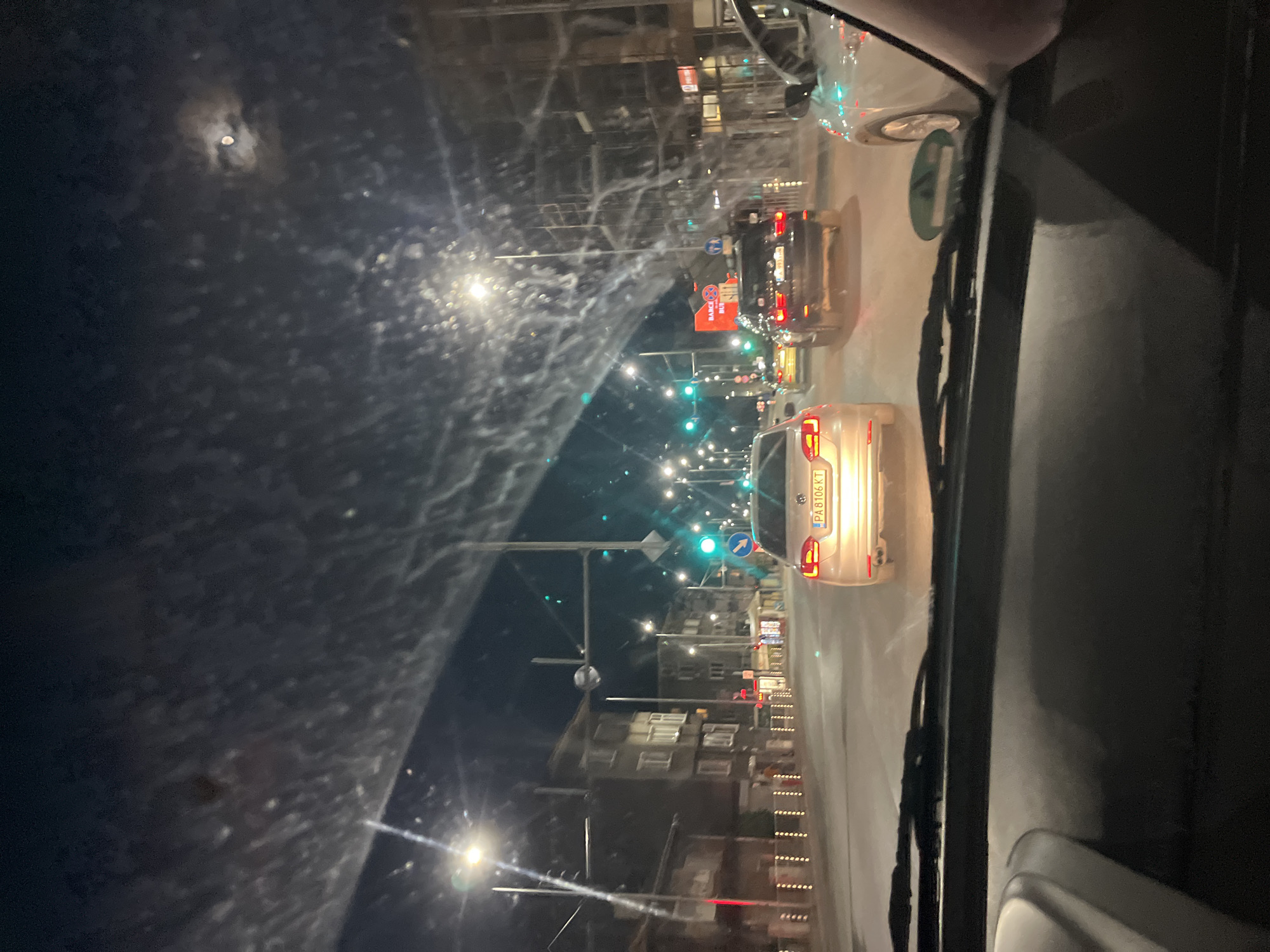 This is a nighttime, interior, point-of-view shot from a vehicle, looking through a dirty windshield at a street scene in Plovdiv, Bulgaria. The windshield is streaked with white smudges, obscuring some of the view. Ahead, a silver Volkswagen sedan with license plate "PA 8106 KT" is visible, followed by a dark SUV. Traffic lights are green, indicating traffic can proceed. Buildings line the street on the left, appearing to be apartments or shops, with lights on inside some of them. Streetlights illuminate the road. A partial view of the moon is visible through the clouds in the dark sky. The interior of the car includes the dashboard and parts of the windshield wipers.
