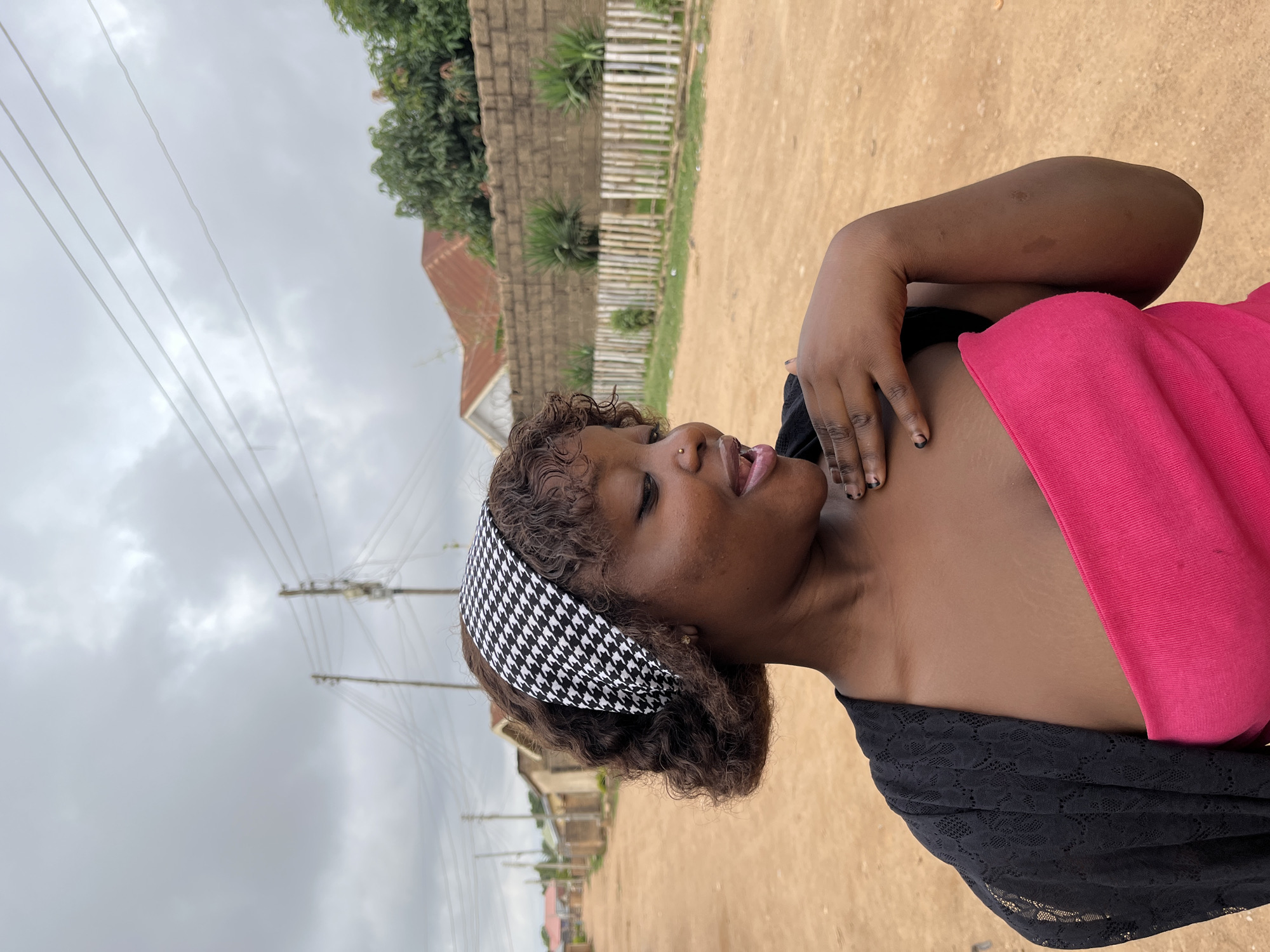 The image, taken in Barakin Akawo, Nigeria, captures a young adult Black woman outdoors on an overcast day. She is positioned on the right side of the frame, looking towards the left with her mouth slightly open, revealing her tongue, suggesting she might be mid-speech or expressing surprise. Her gaze is directed upwards. She has curly, dark brown hair styled with a black and white houndstooth patterned headband. A small gold stud piercing is visible on her right nostril.
She is wearing a black garment, possibly a top or dress, with a textured, lace-like pattern visible on the shoulder/sleeve area. A vibrant pink fabric, possibly a shawl or part of her attire, is draped over her left shoulder and chest. Her right hand is raised towards her chest, displaying dark nail polish on her fingernails.
The setting appears to be a rural or semi-urban environment. The ground is a light brown, unpaved surface, suggesting a dirt road or path. In the background, various structures are visible, including walls and buildings made of reddish-brown bricks, along with some reddish-brown roofs. A fence made of vertical wooden slats is also present. Green trees and shrubbery are scattered throughout the background. The sky is uniformly overcast, filled with light gray clouds, indicating daytime. Utility poles and multiple overhead power or communication lines stretch across the left side of the image against the cloudy sky. No text is visible in the image.