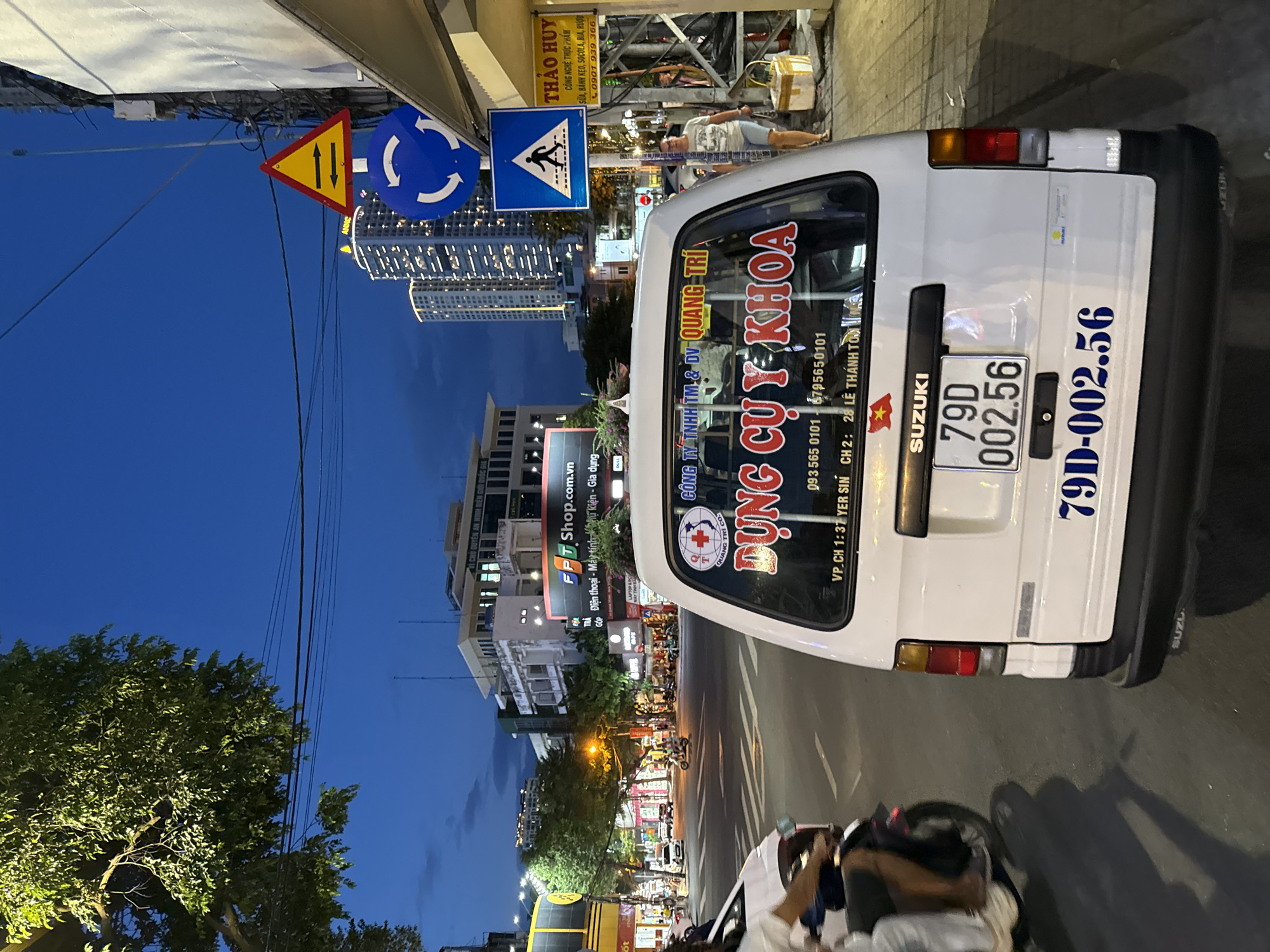 An eye-level, exterior, daylight, medium shot captures a white Suzuki van parked on a city street at dusk. The van faces away from the viewer, with its rear doors open, revealing a glimpse of the interior. A license plate with the characters "79D 002.56" is affixed to the back of the van. The van is adorned with several stickers, one of which features a red and white cross inside a circle with the text "CÔNG TY TNHH TM & DV QUANG TRI" and "DUNG CỤ Y KHOA". Another sticker displays a Vietnamese flag.
The street is paved with asphalt and marked with white lane lines. In the background, a few cars and motorcycles are visible, suggesting moderate traffic. Buildings line the street, including a multi-story structure with visible signage and illuminated windows. A few streetlights cast a warm glow on the scene. The sky is a deep blue, indicating the transition from day to night. Pedestrians are present on the sidewalk, and a traffic light is visible in the distance. The overall atmosphere is that of a busy urban environment during the evening hours. The image is taken in Nha Trang, Vietnam.