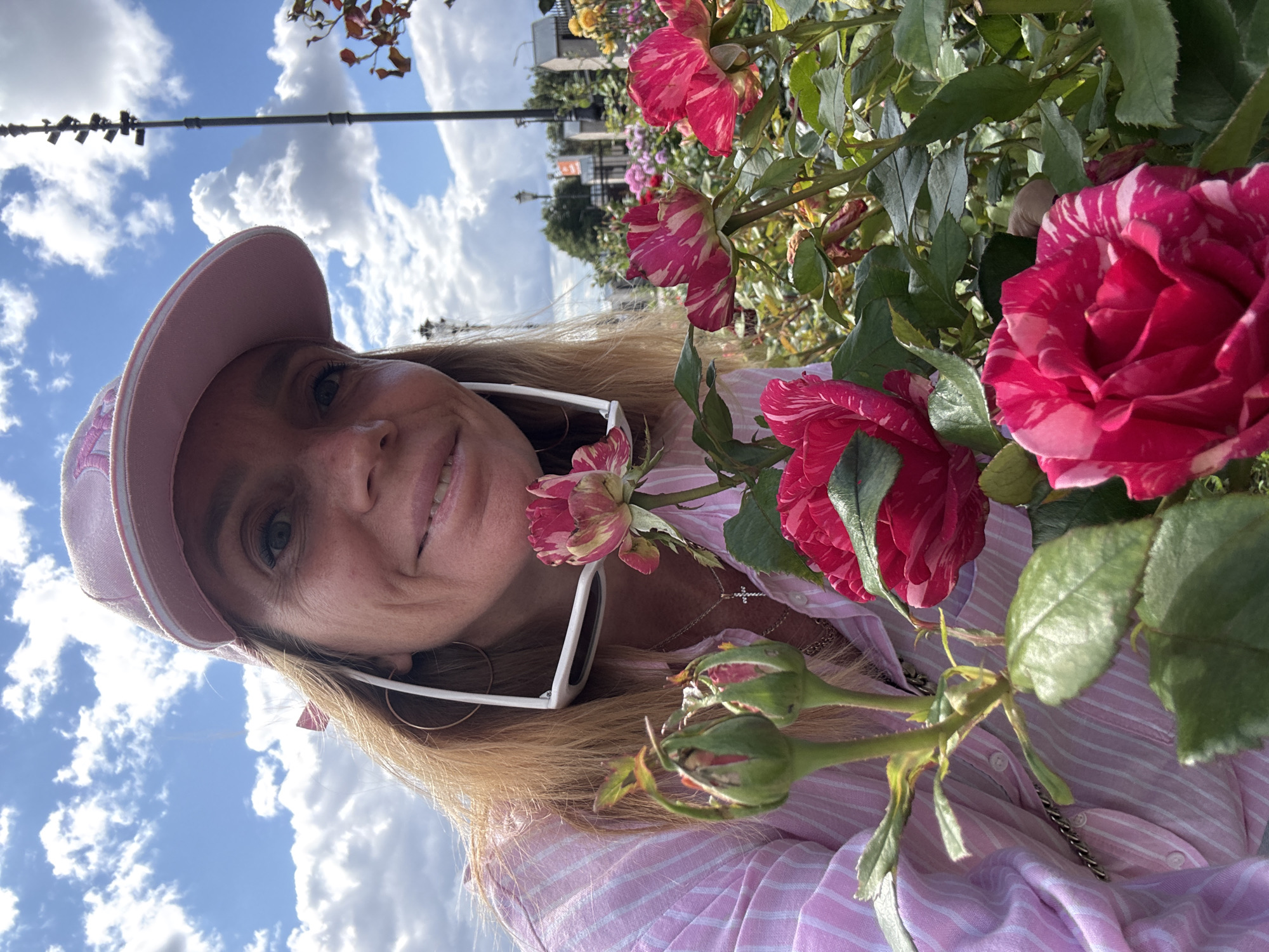 A woman with blonde hair, wearing a pink baseball cap and a pink striped shirt, smiles at the camera. She has sunglasses pushed up on her head. She is surrounded by blooming pink and white striped roses. The background shows a cloudy blue sky, a tall pole with lights, and a garden with various flowers and trees. The scene appears to be outdoors in a park or botanical garden.