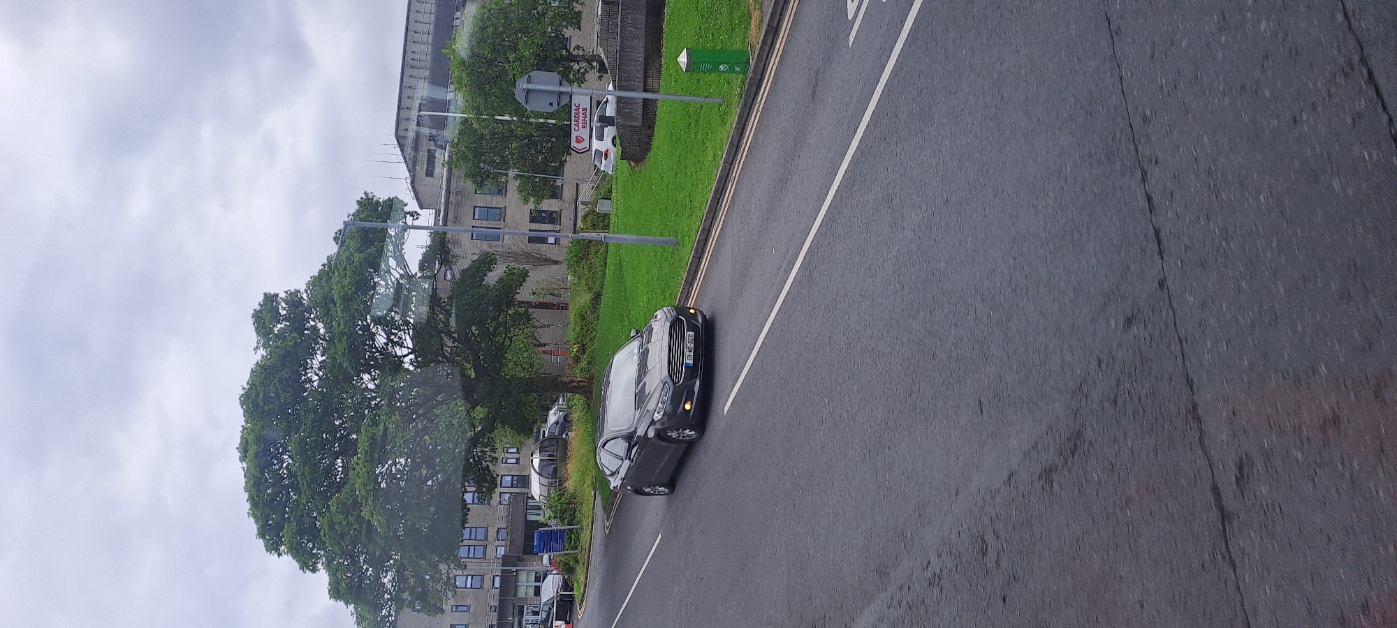 The image shows a dark gray Ford sedan driving on a wet road in what appears to be a hospital or medical facility campus. The car has the license plate "171-WD-3072". In the background, there is a large, mature tree with dense foliage, and a multi-story building with a grey facade and numerous windows. A sign with a red heart icon and the text "CARDIAC REHAB" is visible on the right side of the road, indicating the location is likely associated with cardiac rehabilitation services within a hospital. The sky is overcast and the road surface shows signs of rain.