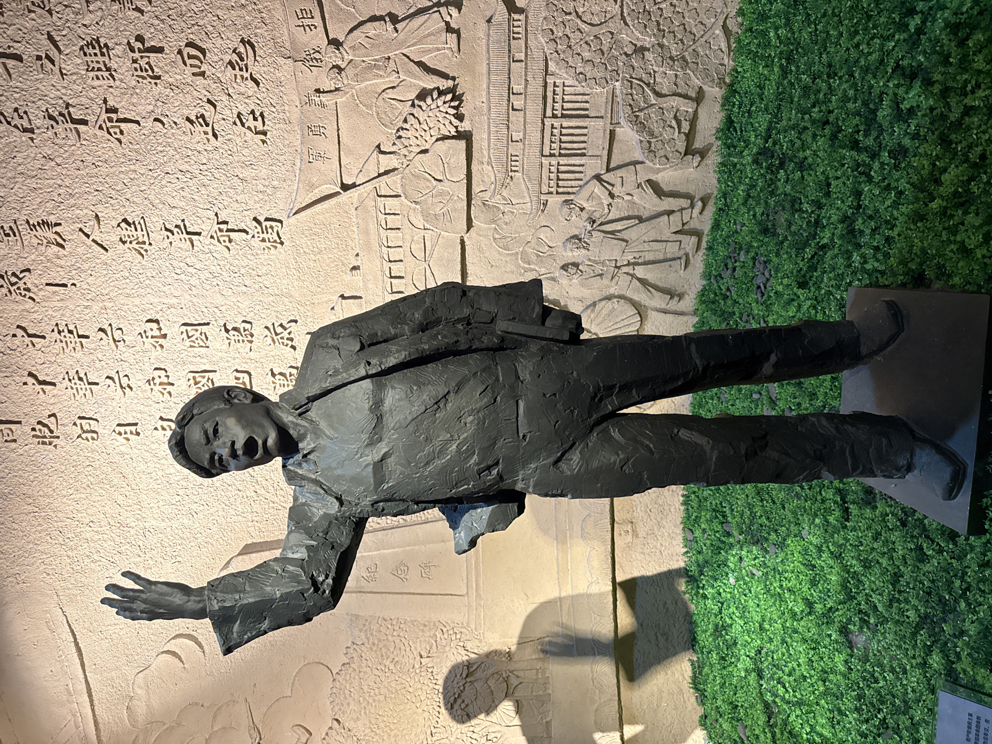 The image shows a statue of a man in a suit with his mouth open and arm extended. Behind the statue is a wall with carvings and Chinese characters. To the right is a wall of green foliage. The statue is located in the Yuzhong District of China.