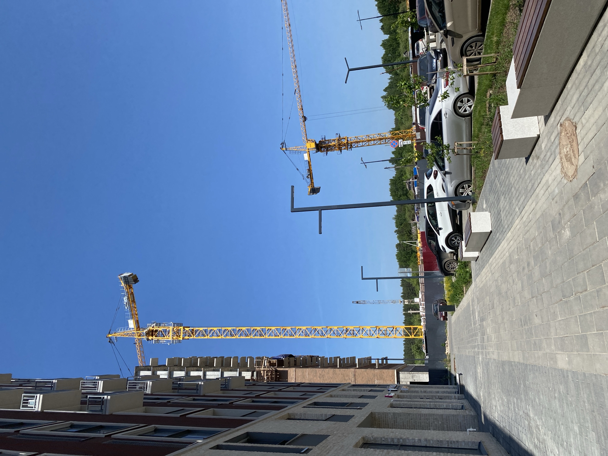 The image shows a construction site in Izhevsk, Russia. A building under construction occupies the lower left of the frame, with two yellow tower cranes looming above it against a clear blue sky. Along the right side of the image, a gray brick walkway stretches into the distance, lined with parked cars, streetlights, benches, and young trees. The presence of construction and parked vehicles suggests an urban area undergoing development.