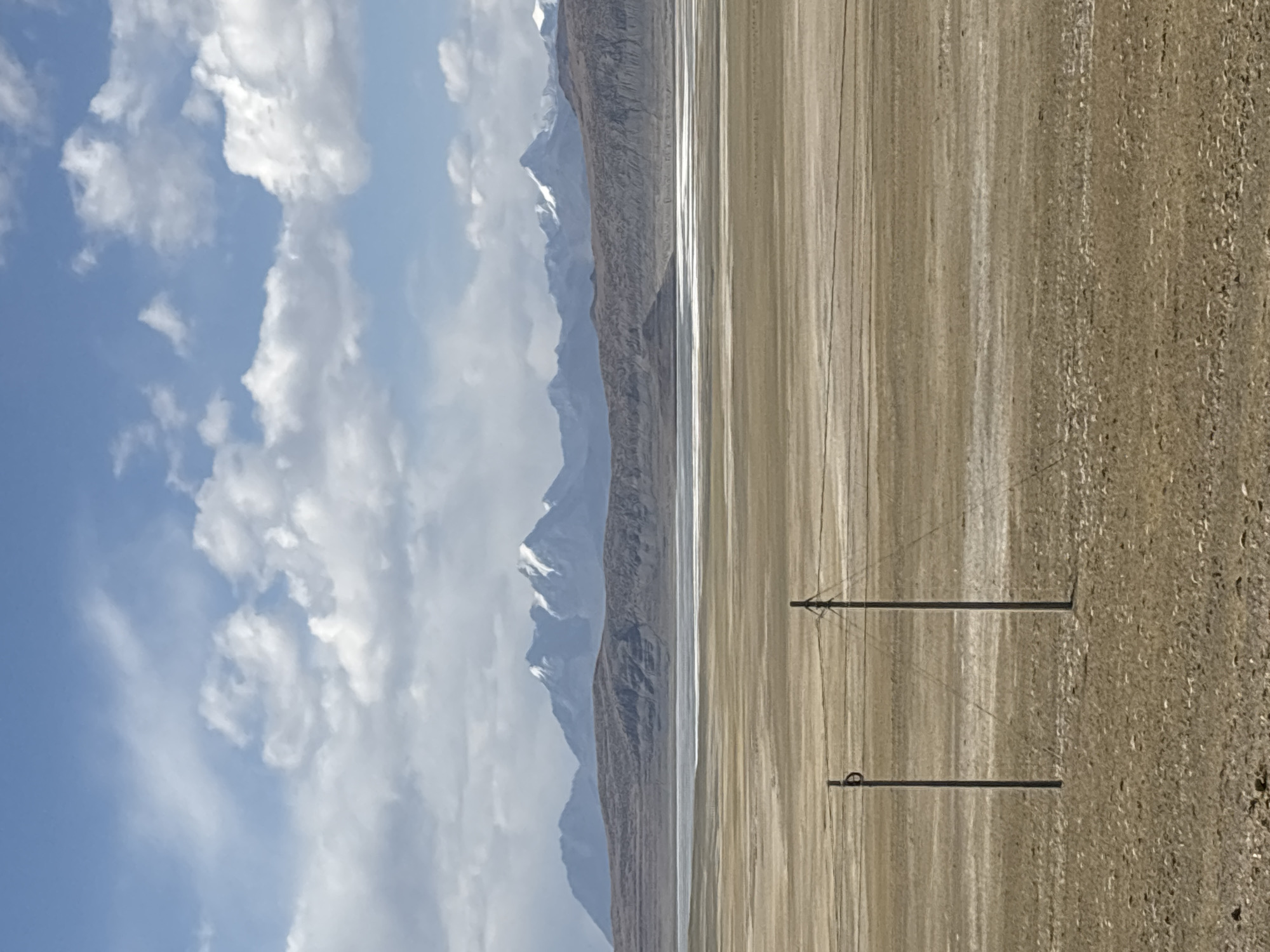The image shows a horizontal landscape in Gyirong County, China. A partly cloudy, blue sky occupies the left half of the frame. Below the sky and slightly right of center, a series of tall, rugged mountains stretch along the horizon with snow visible on their peaks.
A flat, beige-colored plain extends across the middle and right portions of the image. Two utility poles are visible in the foreground. The plain appears to have a slight reflective surface, suggesting it could be a salt flat or a shallow body of water. The ground transitions from a smoother, light-colored texture near the mountains to a coarser, darker texture towards the right.