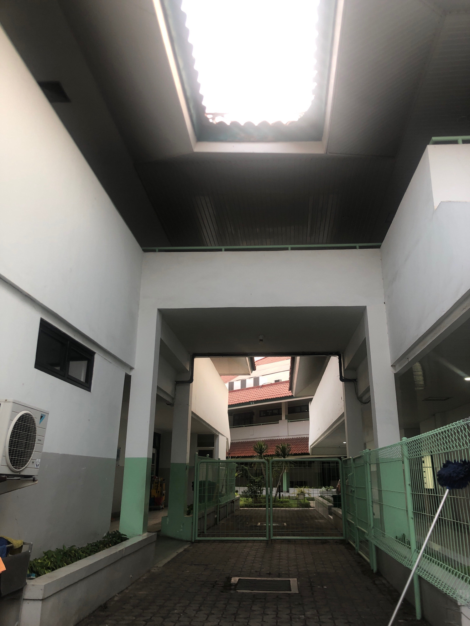 The image captures a view of an outdoor walkway or corridor, possibly part of a school or residential complex, with a skylight at the top allowing natural light to filter through. The architecture features white walls and a dark, slatted ceiling. On the left, a white air conditioning unit is mounted on the wall, below a dark-framed window. The walkway leads to an open area with a green metal gate. In the background, a multi-story building with red-tiled roofs is visible. The ground is paved with interlocking bricks. The overall scene suggests an urban environment in Surabaya, Indonesia, characterized by its functional architecture and typical weather patterns of the region.