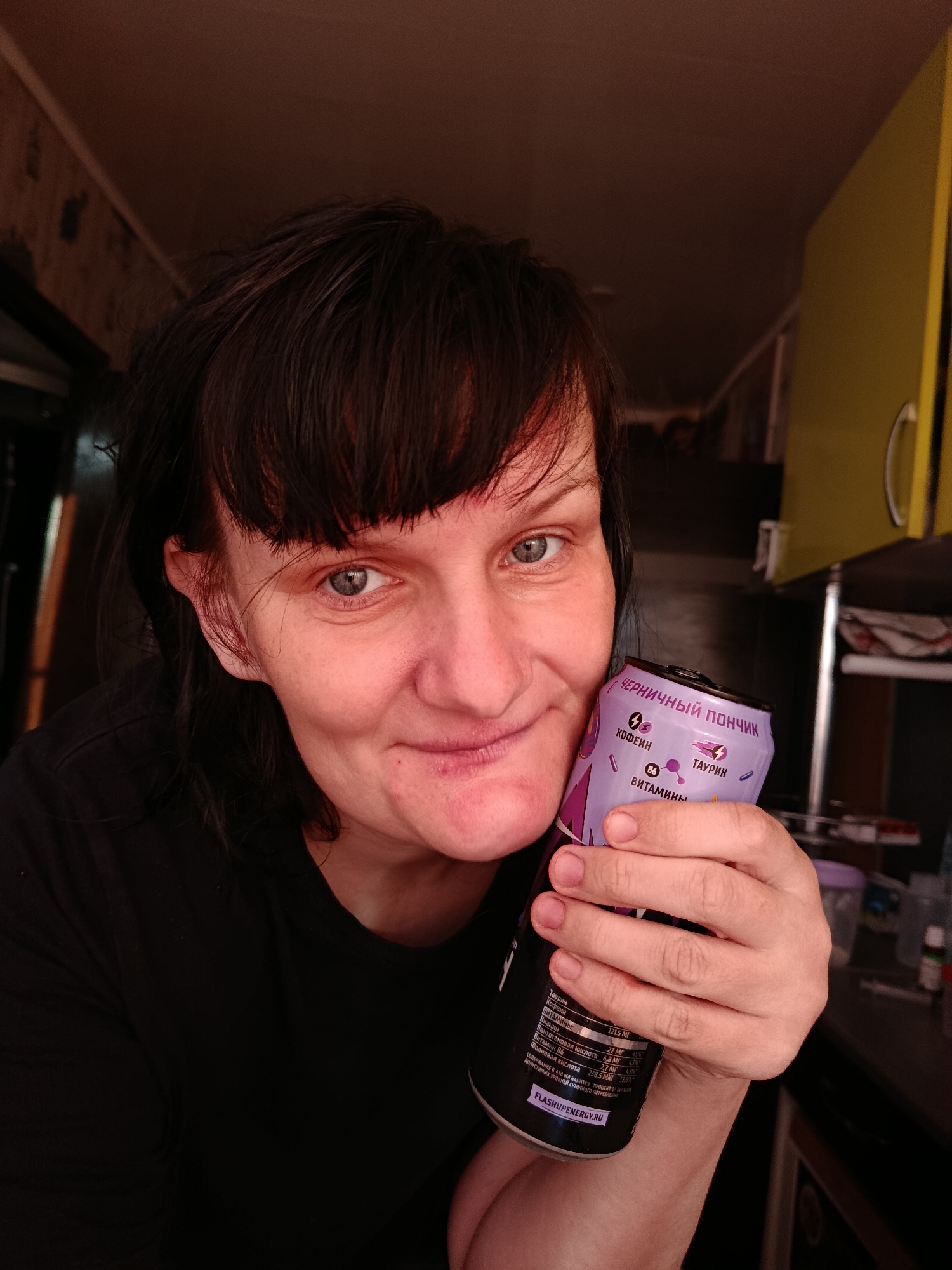 A close-up selfie shows a woman with dark, medium-length hair and bangs, looking directly at the camera with a slight, gentle smile. Her light-colored eyes are prominent. She is wearing a black t-shirt. She holds a purple and black energy drink can with her right hand, positioned close to her face.
The can's label is primarily in Russian, with "ЧЕРНИЧНЫЙ ПОНЧИК" (Blueberry Donut) clearly visible at the top, likely indicating the flavor. Other prominent words on the can include "КОФЕИН" (Caffeine), "ТАУРИН" (Taurine), and "В6 ВИТАМИНЫ" (B6 Vitamins). Detailed nutritional information is also legible, listing components like "Таурин," "Кофеин," "Ниацин," "Пантотеновая кислота," "Витамин В6," and "Фолиевая кислота," along with their respective values and percentage daily allowances. The website "FLASHUPENERGY.RU" is visible at the bottom of the can.
The background suggests an indoor setting, possibly a kitchen or a room with kitchen elements. A muted, textured wallpaper is visible on the left wall, while a bright yellow cabinet or shelf is seen on the right. Below the yellow cabinet, a darker counter or shelf holds various small objects, possibly bottles or containers. The lighting is soft, suggesting an indoor environment without strong direct light. The woman's expression is friendly and relaxed.
