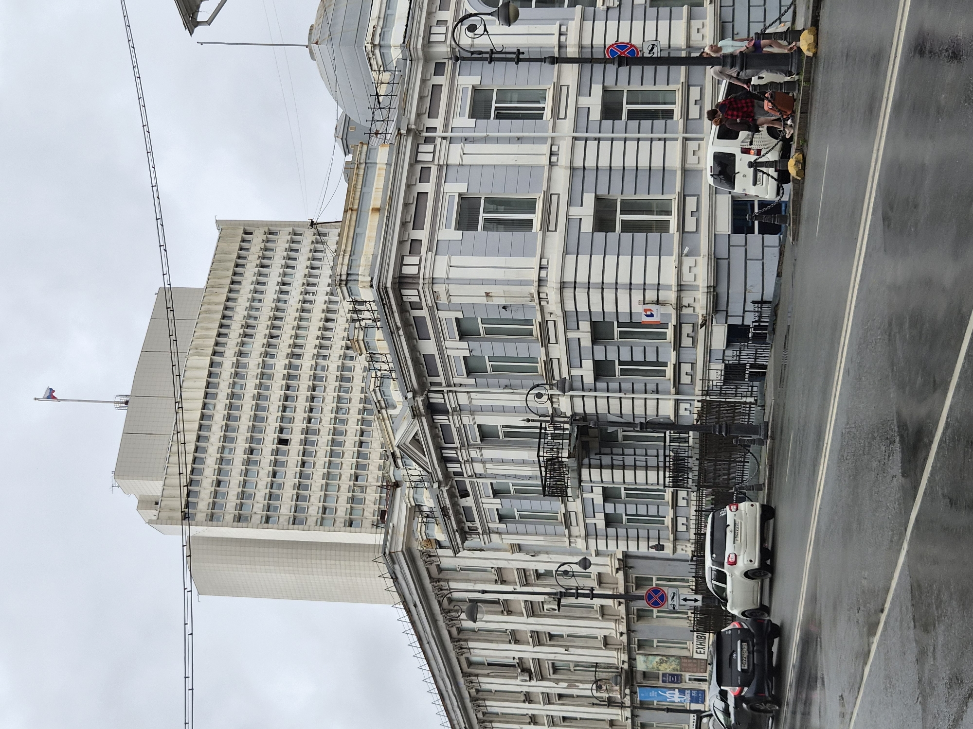 The image captures a street scene in Vladivostok, Russia, dominated by a grand, old European-style building with light blue and white facade, featuring intricate architectural details such as columns, decorative moldings, and wrought-iron balconies. Behind this building, a much larger, modern, beige high-rise structure stands prominently against an overcast sky. The street in the foreground is wet, indicating recent rain, with white lane markings visible. Several cars are parked or driving on the street. To the right, a white van is parked, and three people are gathered near it, one of whom is handling luggage. Another person is standing next to the van, and a third is walking away. A traffic sign with a red circle and diagonal bar is visible on a lamppost. The overall atmosphere is somber, likely due to the cloudy weather.