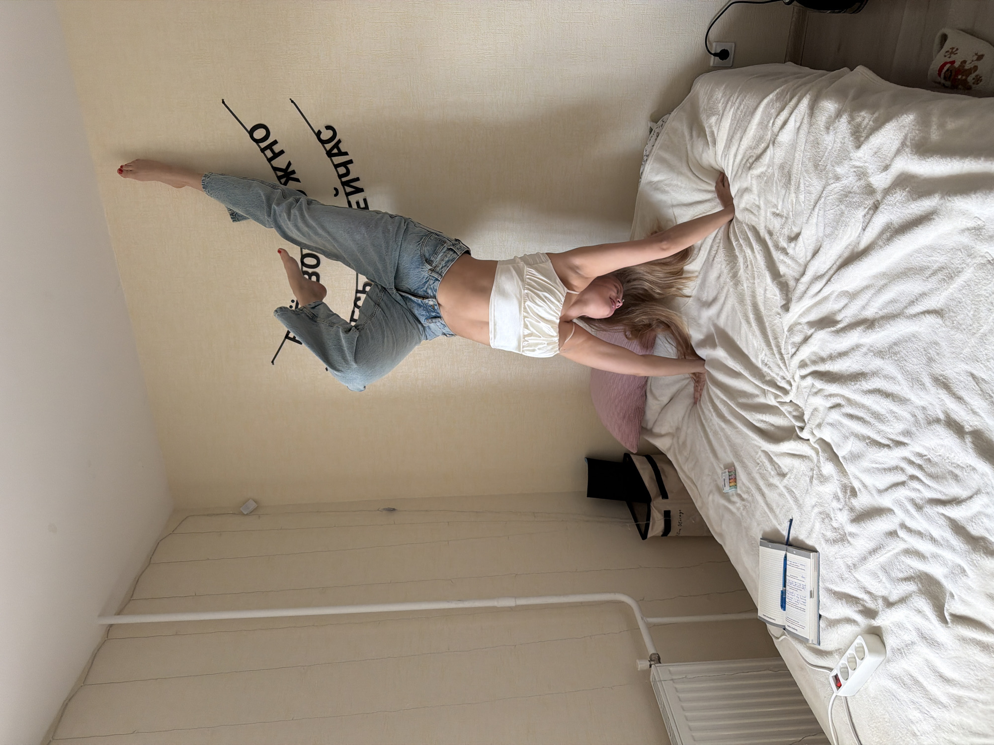 A young woman wearing jeans and a white top lies on a bed. She is positioned diagonally across the frame, with her upper body and head near the top right corner of the bed, and her legs extending towards the upper left corner. A bedside table is located on the right side of the bed. The location is Izhevsk, Russia. The bedroom has a wall-to-wall carpet.