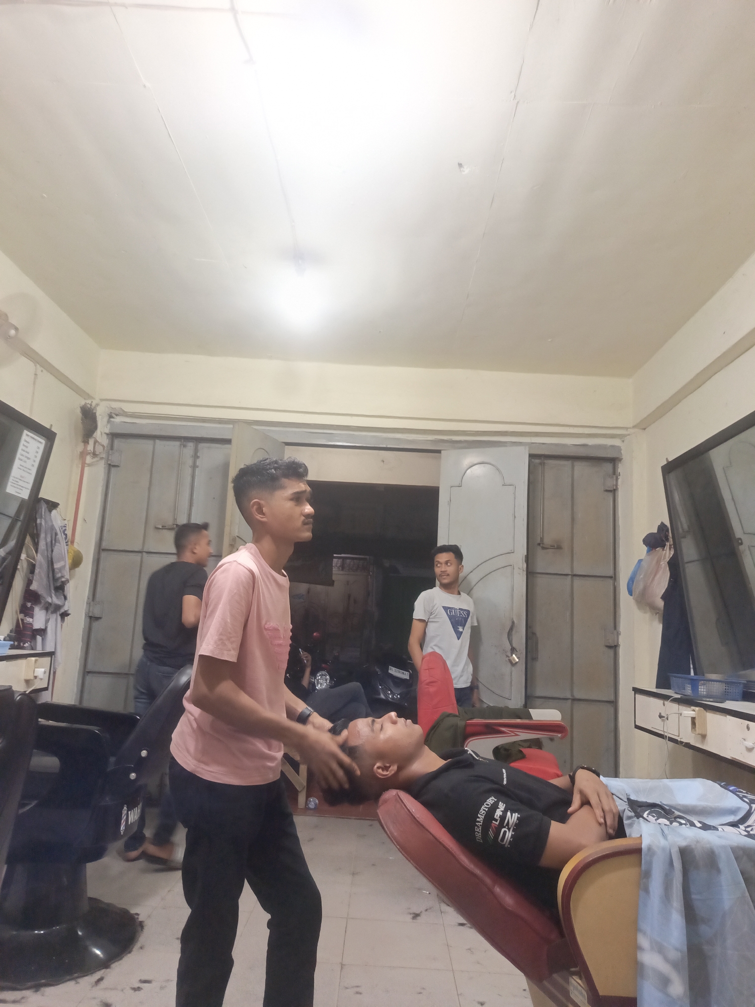 This image depicts the interior of a barber shop in Lhokseumawe, Indonesia. The scene is illuminated by overhead lighting, casting a warm glow on the beige-painted walls and ceiling. In the foreground, a young man in a light pink t-shirt and black pants is standing and attending to another young man who is reclining in a red barber chair. The seated man is wearing a black t-shirt and has his eyes closed, appearing to be receiving a head massage or hair treatment. A light blue and white patterned drape is laid across his lap and the chair. To the left, a black barber chair with the word "WAR" visible on its base is partially visible. Further back, two other young men are present. One is standing in a grey t-shirt with a "GUESS" logo, looking towards the right. Another man in a black t-shirt and jeans stands near a large, grey metal door at the back of the shop. The environment suggests a typical barbershop setting with some evidence of recent activity, such as scattered hair clippings on the tiled floor.