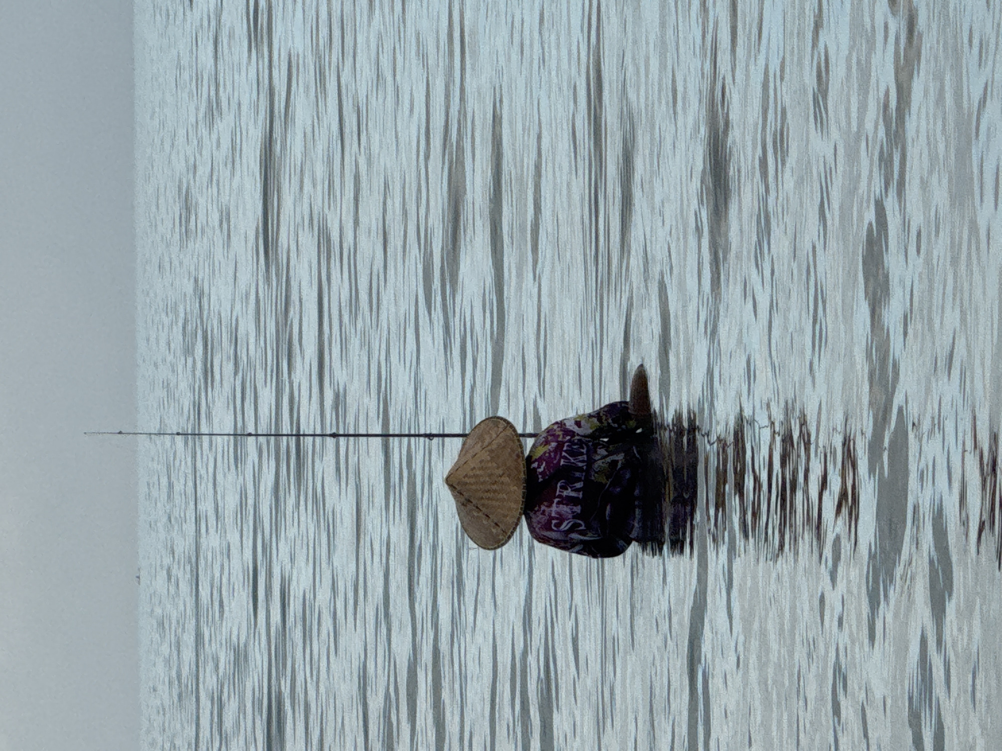 A person wearing a conical hat is fishing in water. The water has a rippled surface. The location is Kuta Selatan, Indonesia. A fishing rod extends into the water. The person is wearing a shirt with a word on it.