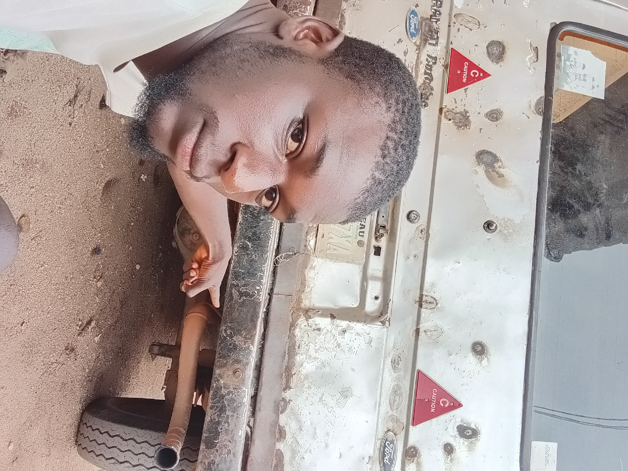 The image depicts a man pointing to the exhaust pipe of a weathered vehicle, possibly in Bukuru, Nigeria. The vehicle's rear is visible, showing signs of rust and damage. Two red triangular "CAUTION" stickers are affixed to the vehicle's body, one on the left and one on the right. A faded "Ford" logo is present on the left side of the vehicle's rear. A license plate with the inscription "EAU" and "XA" is partially visible. The ground is covered in sand and small debris. The man is in the foreground, looking towards the camera. His right hand is extended, with his index finger pointing towards the exhaust system.