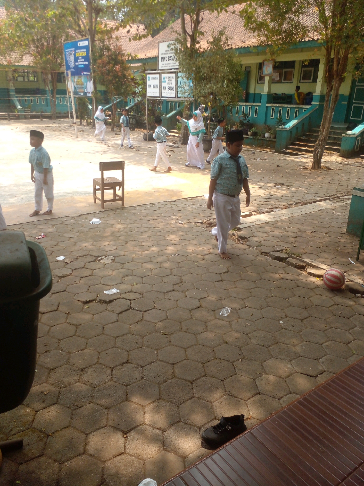 The image shows an outdoor scene at a school in Banyuputih, Indonesia, likely during daylight hours due to the strong shadows. Several children in school uniforms, consisting of light-colored shirts and white pants, are visible. One boy in the foreground wears a patterned shirt and tie, along with white pants and a black peci (cap). Another boy in a similar uniform stands to his left. Further in the background, other children are seen walking. The school building, painted in light green and turquoise, features a prominent tiled roof. The ground is paved with hexagonal concrete blocks, and there is scattered litter on the ground, including plastic wrappers and paper. A wooden chair is placed on the pavement. A red and white striped ball lies on the ground to the right, and a black shoe is visible on a brown wooden bench in the lower right corner. Trees with green foliage are present, suggesting a well-maintained outdoor area. Two large signs with text in Indonesian are visible on poles in the background, indicating this is an educational institution.