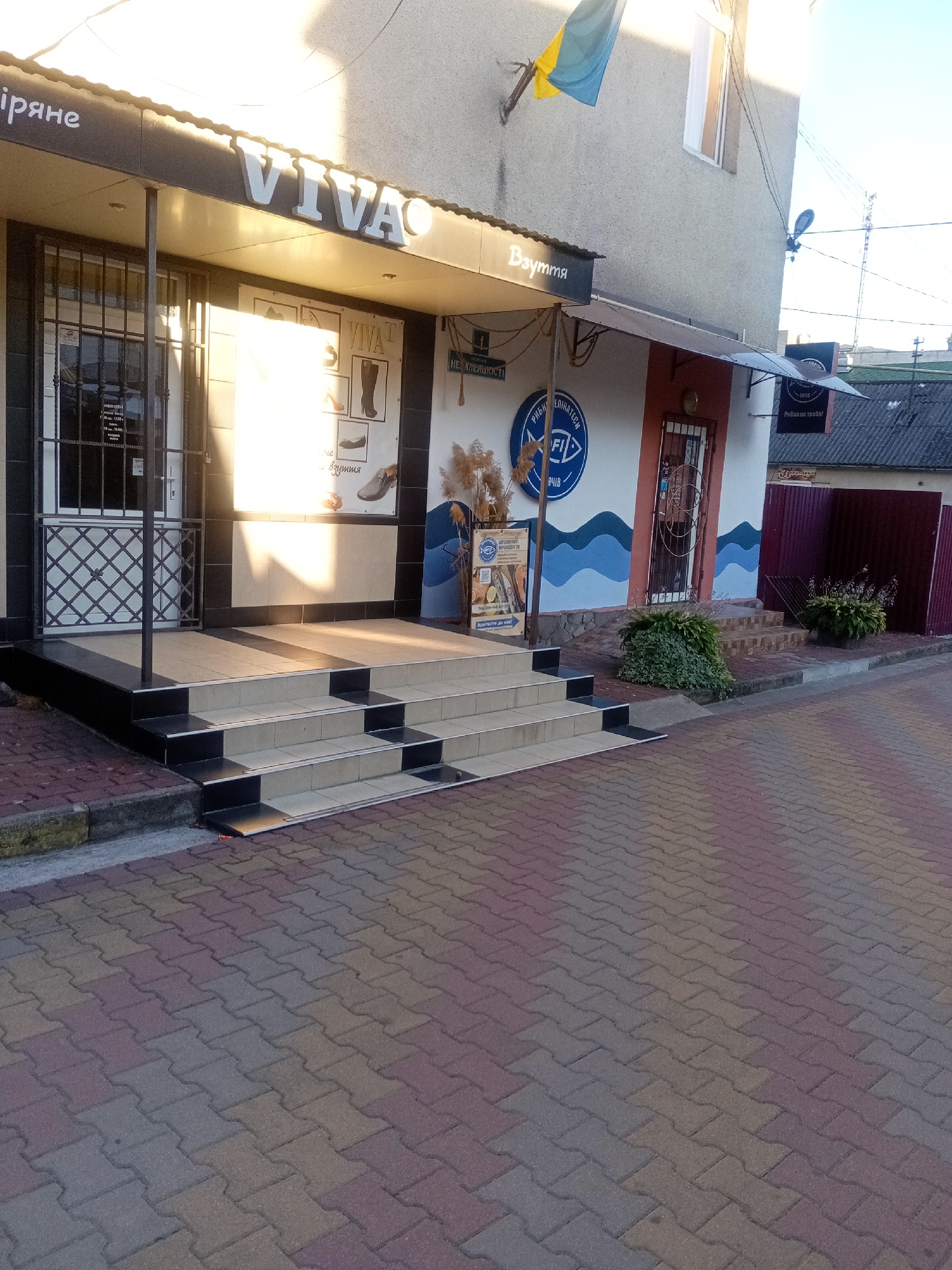 The image shows the exterior of a store named "VIVA" in Tiachiv, Ukraine, with a sign that reads "Взуття" (Footwear). The store has a modern facade with large windows and a covered entrance. A Ukrainian flag is visible flying from the building. The entrance is accessed by a set of tiered steps made of beige and black tiles. The ground in front of the store is paved with interlocking bricks in shades of red, grey, and yellow. To the right of the entrance, there is a mural depicting stylized waves and a logo for a fishing store. There are potted plants and bushes near the entrance and along the sidewalk. The street appears to be a commercial area with other buildings visible in the background. The lighting suggests it is daytime, possibly late afternoon, with sunlight casting shadows on the pavement and building. There are no people visible in the image.