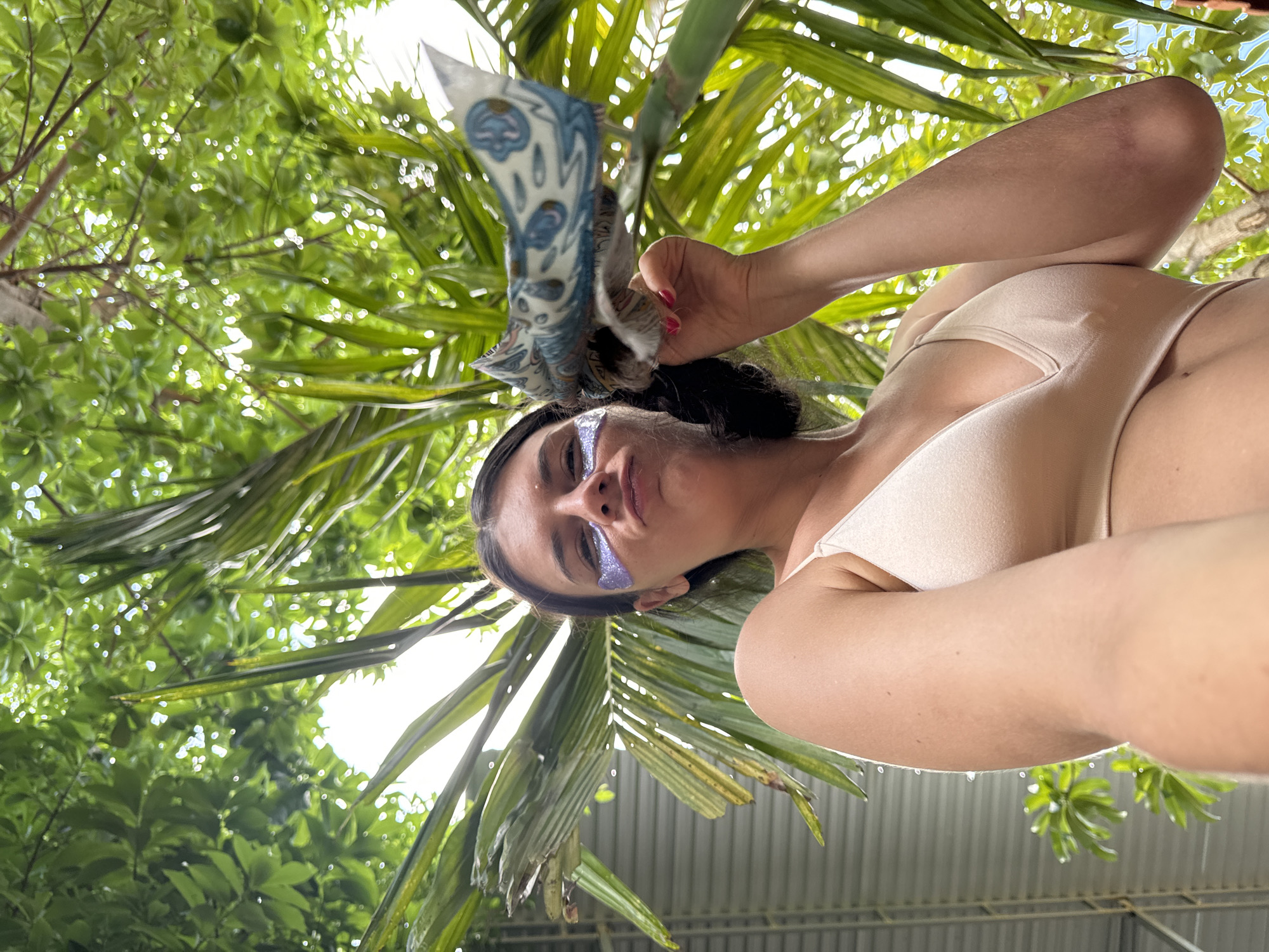 A person with dark hair and glitter on their face is shown from a low angle, looking up at the camera. They are wearing a light-colored bikini top and holding a blue and white patterned fabric to their forehead. The background is filled with lush green foliage, including palm fronds, and hints of sunlight filtering through the leaves. The overall scene appears to be outdoors, possibly in a tropical or subtropical environment. The lighting suggests it is daytime.