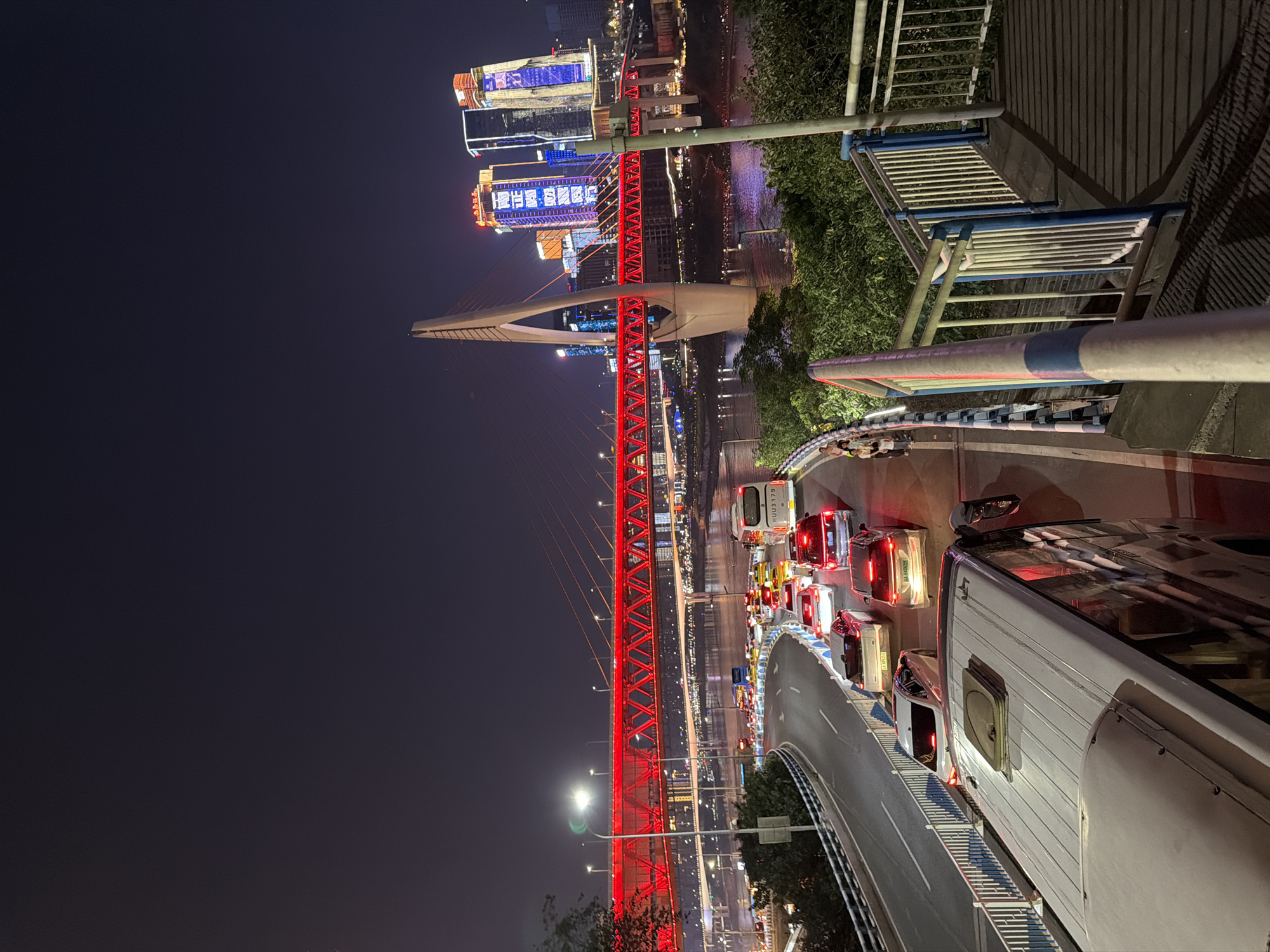 The image depicts a nighttime view of a busy highway intersection with a river and illuminated skyscrapers in the background, characteristic of Yuzhong District, China. A prominent red suspension bridge spans the river, its structure outlined with bright lights. The foreground features a multi-lane road filled with numerous vehicles, including cars, taxis, and buses, indicating heavy traffic. Several cars are stopped, with their brake lights illuminated, suggesting a traffic jam. A white van is parked on the right side of the road in the foreground. People are visible walking on a sidewalk to the left, alongside the road. Lush green trees and bushes line the riverbank and the sides of the elevated roadway. The cityscape is dominated by modern skyscrapers, brightly lit with colorful LED displays. The sky is dark, typical of nighttime.