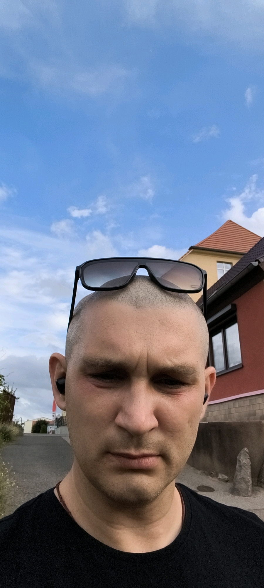A man with a shaved head and sunglasses perched on top of his head, wearing a black t-shirt and black earbuds, is looking directly at the camera. He has a stern expression on his face, with his brow furrowed. Behind him, a red building with a peaked roof is visible, along with a street and some greenery. The sky is blue with scattered clouds. The man is standing on a paved surface in what appears to be a residential area. The scene is likely captured in Wolgast, Germany, given the architectural style of the buildings.