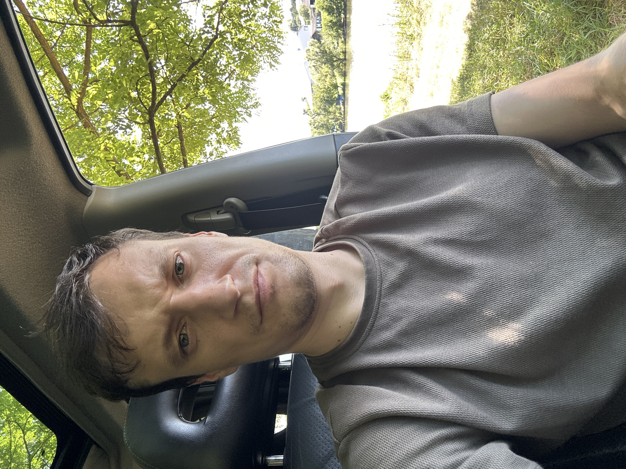 A man is taking a selfie in a car with a sunroof open. Sunlight filters through green leaves overhead, casting shadows on the car's interior and the man's face. The man has short brown hair, fair skin, and a neutral expression. He is wearing a brown t-shirt. Through the car window, a body of water with trees and buildings on the opposite bank is visible. The setting appears to be a natural environment near a body of water. The specific location is Makiv, Ukraine.
