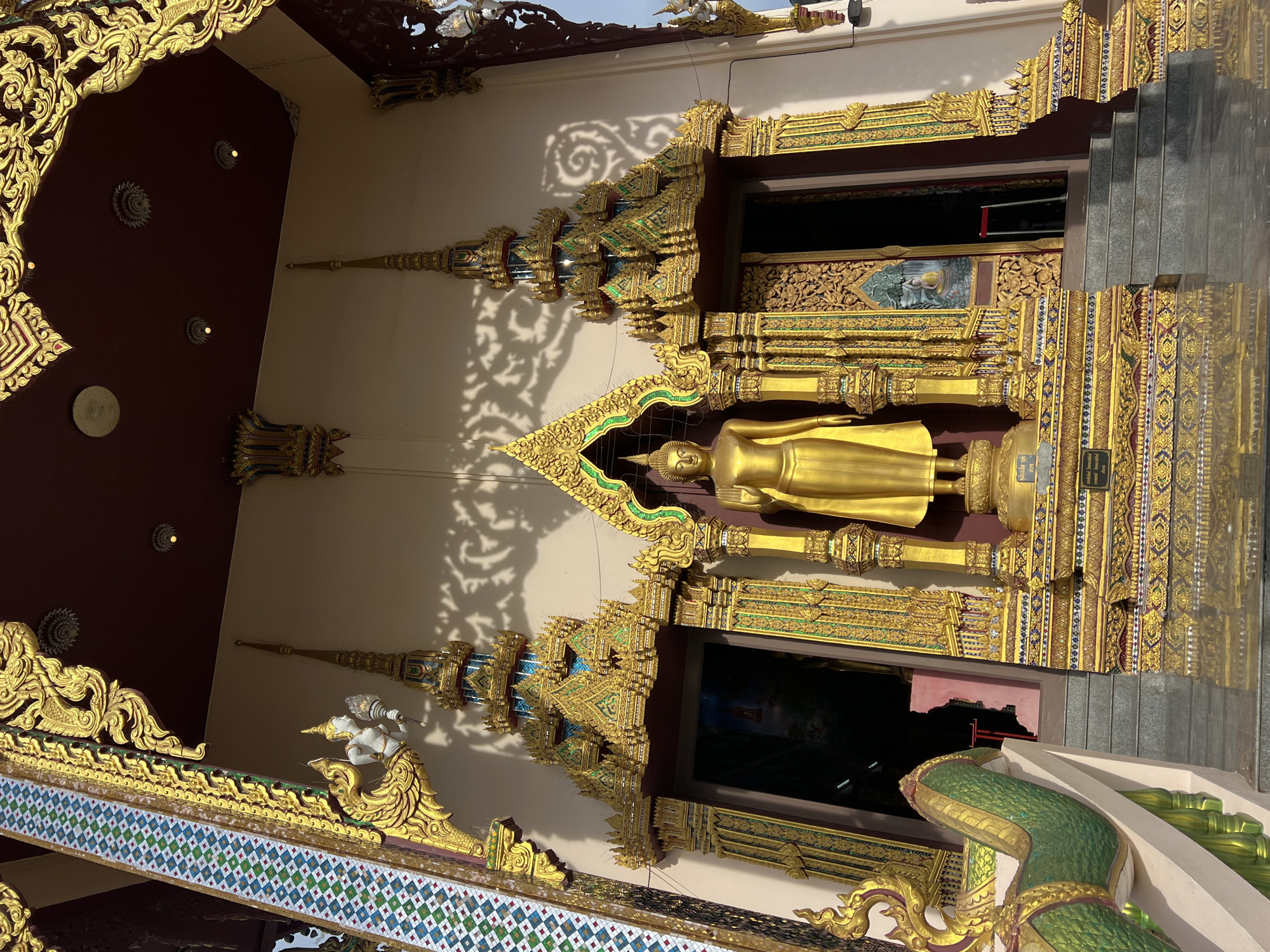 The image shows the entrance to a temple in Baan Plai Laem, Thailand. A large, golden statue of the Buddha stands prominently in the center, flanked by ornate, multi-tiered golden structures with colorful detailing. To the left and right of the central entrance are doorways leading into the temple, also adorned with elaborate golden carvings. A staircase leads up to the entrance. The architecture features intricate golden scrollwork, mythical creature motifs, and vibrant mosaic tiles. The building's facade is a light beige color, with shadows from overhead structures casting decorative patterns on the wall. A green serpent-like sculpture is visible on the left side of the image, forming part of the temple's exterior decoration. The sky is visible in the upper right corner, indicating an outdoor scene.