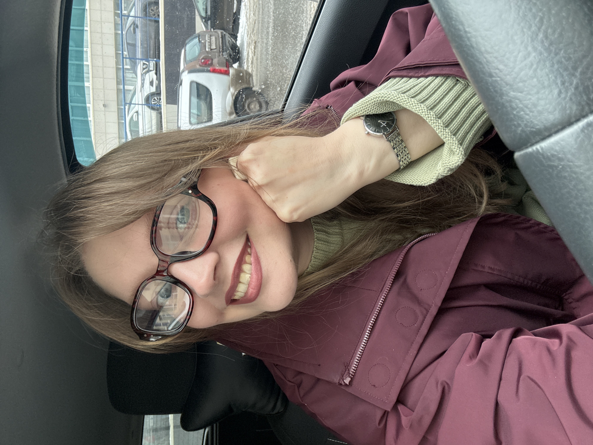 A woman is in a car, smiling and looking at the camera. She is wearing glasses, a green sweater, a maroon jacket, and a watch. The background is blurry, but it appears to be an urban environment. Outside, there are other cars, buildings with blue and white siding, and overcast skies. The location is Izhevsk, Russia.
