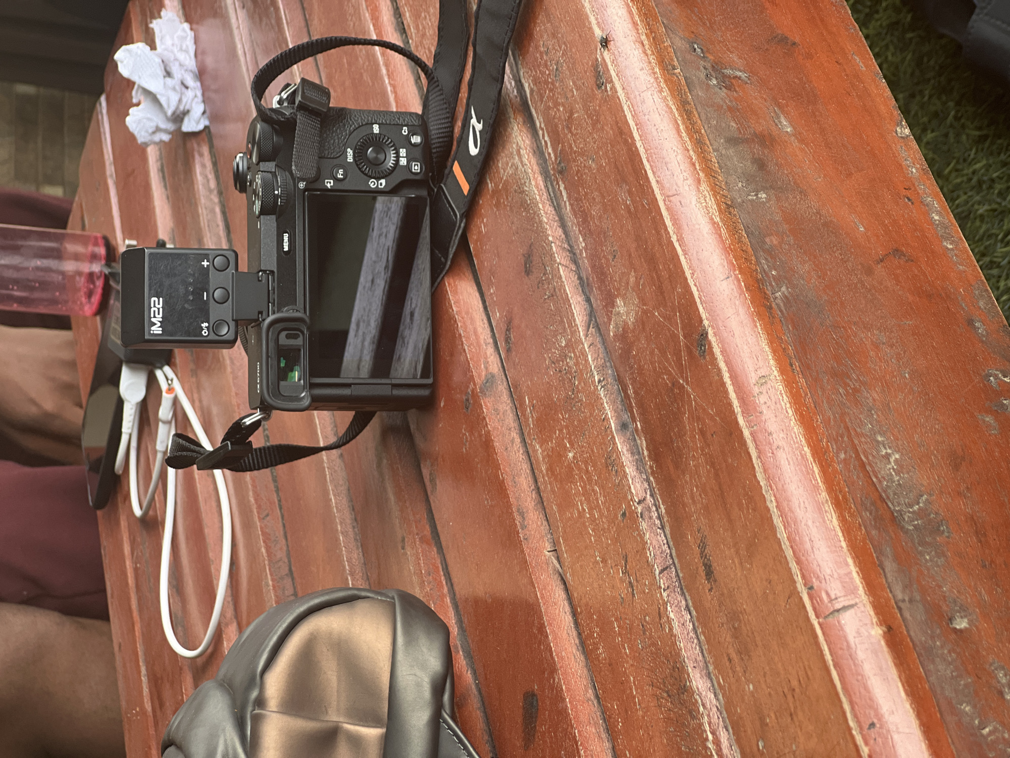 An overhead shot captures a collection of electronic devices and personal items resting on a rustic wooden surface, likely an outdoor table or bench. The central subject is a black digital camera, identifiable as a Sony Alpha series due to the "α" logo visible on its black strap. The camera's screen is flipped out and angled, reflecting diagonal lines from the wooden planks.
Attached to the camera's hot shoe is a compact black accessory, possibly an external microphone or audio recorder. This device prominently displays the text "iM22" and features three buttons: a "+" for increasing, a "-" for decreasing, and a circular button at the bottom that appears to be a power or mode control, with what looks like "O/C" or "O/G" printed next to it. Several buttons and dials are visible on the camera's top and rear, including "DISP", "Fn", "ISO", "MENU", and icons for playback, trash, and wireless connectivity.
To the left of the camera, a white power bank is visible, connected by white USB cables to a dark smartphone (partially obscured) and potentially other devices. A red transparent water bottle is also present nearby. A piece of crumpled white paper or tissue lies further left on the wood. A partial view of a dark bag or backpack is seen in the lower foreground.
The wooden surface itself is a prominent feature, composed of wide planks with a reddish-brown hue. It shows signs of wear, including scratches, discoloration, and a somewhat distressed texture. In the background, green grass is discernible, confirming an outdoor setting. The bright, even lighting suggests it is daytime. A person's leg and arm are partially visible in the bottom left, indicating human presence. The overall scene suggests a break or preparation during an outdoor activity, possibly related to photography or content creation.