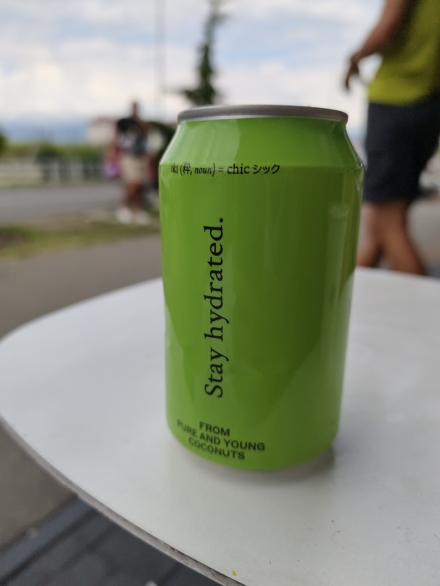 A bright green aluminum can with black text sits on a white table outdoors. The text on the can reads, "iki (粋, noun) = chic シック" and "Stay hydrated." Below this, smaller text says, "FROM PURE AND YOUNG COCONUTS." In the background, a blurred person in a neon yellow shirt and dark shorts is walking away from the camera. The environment appears to be a street or park setting with trees and buildings visible in the distance. The sky is cloudy. The scene suggests a casual outdoor setting, possibly a cafe or public space.