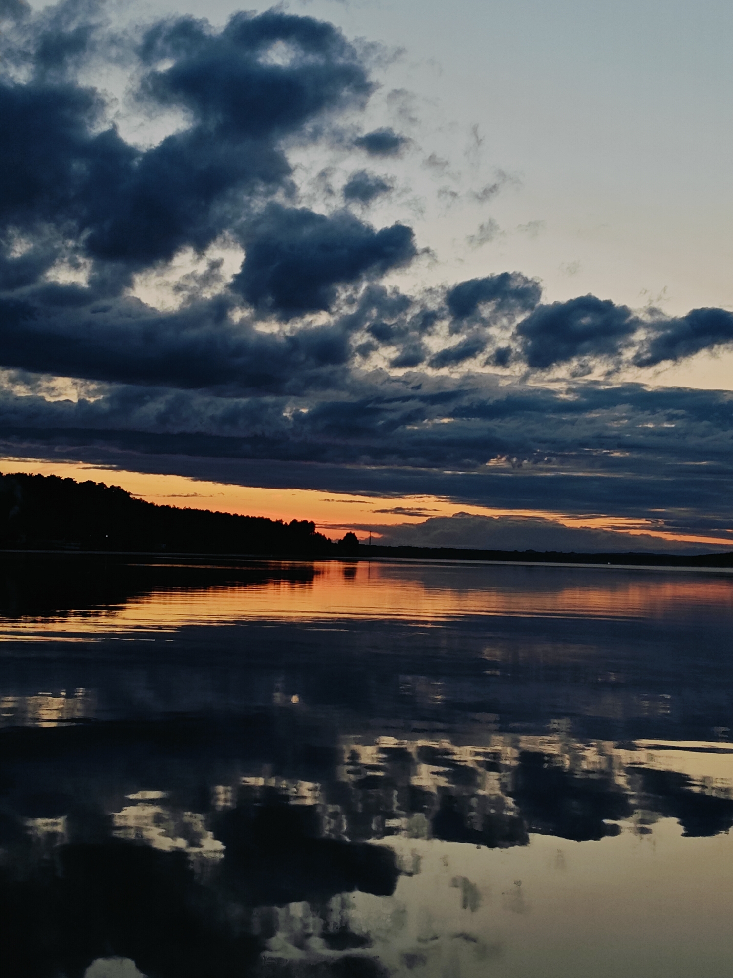 The image depicts a twilight scene over a body of water, likely a reservoir given the context of Chaykovsky, Russia. A silhouetted treeline forms the horizon on the left side of the frame. The sky is filled with dark, dramatic clouds, with streaks of orange and yellow light from the setting sun visible near the horizon, suggesting a sunset or the period just after. The water's surface reflects the sky and the treeline, creating a mirror image. The overall impression is one of natural beauty and tranquility.