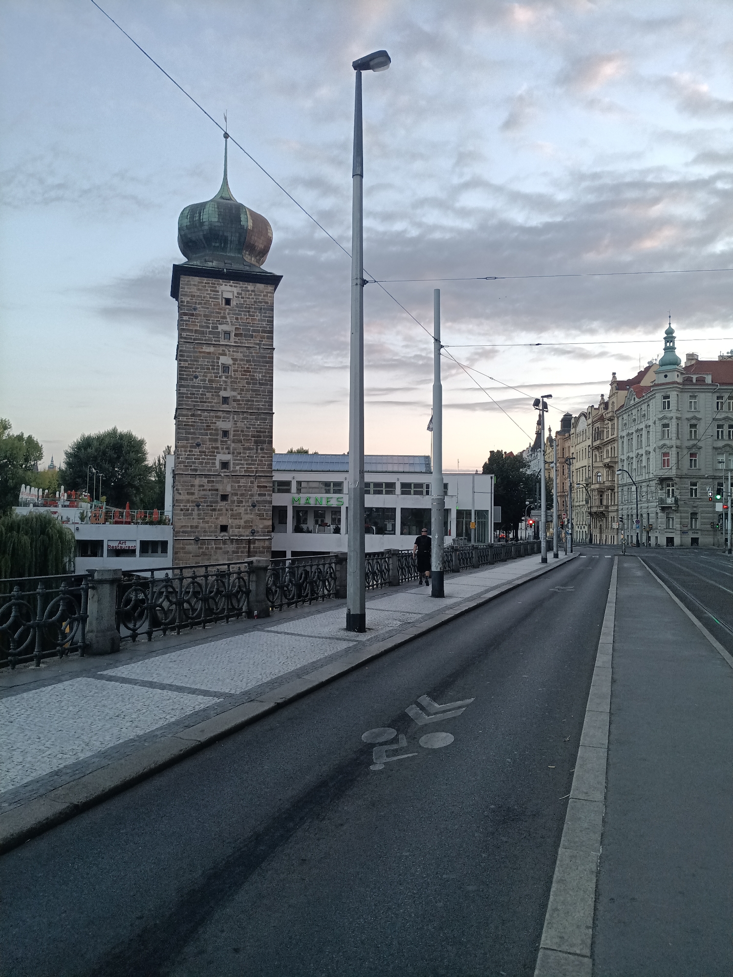 The image depicts a view of the Vltava River embankment in Prague, Czechia, looking east. In the foreground, a paved road with a bicycle lane marking runs parallel to a sidewalk bordered by a decorative metal fence. Across the sidewalk, a stone tower with a green, onion-shaped dome stands prominently, a portion of the "Mánes" building is visible behind it. To the right of the tower, a modern white building with green lettering "Mánes" is situated. Further in the background, a street lined with multi-story buildings with ornate facades stretches into the distance. The sky is overcast, suggesting either early morning or late evening light. A single person, a male wearing dark clothing, is walking on the sidewalk near the railing. The scene is a blend of historic architecture and modern infrastructure, characteristic of Prague's urban landscape.
