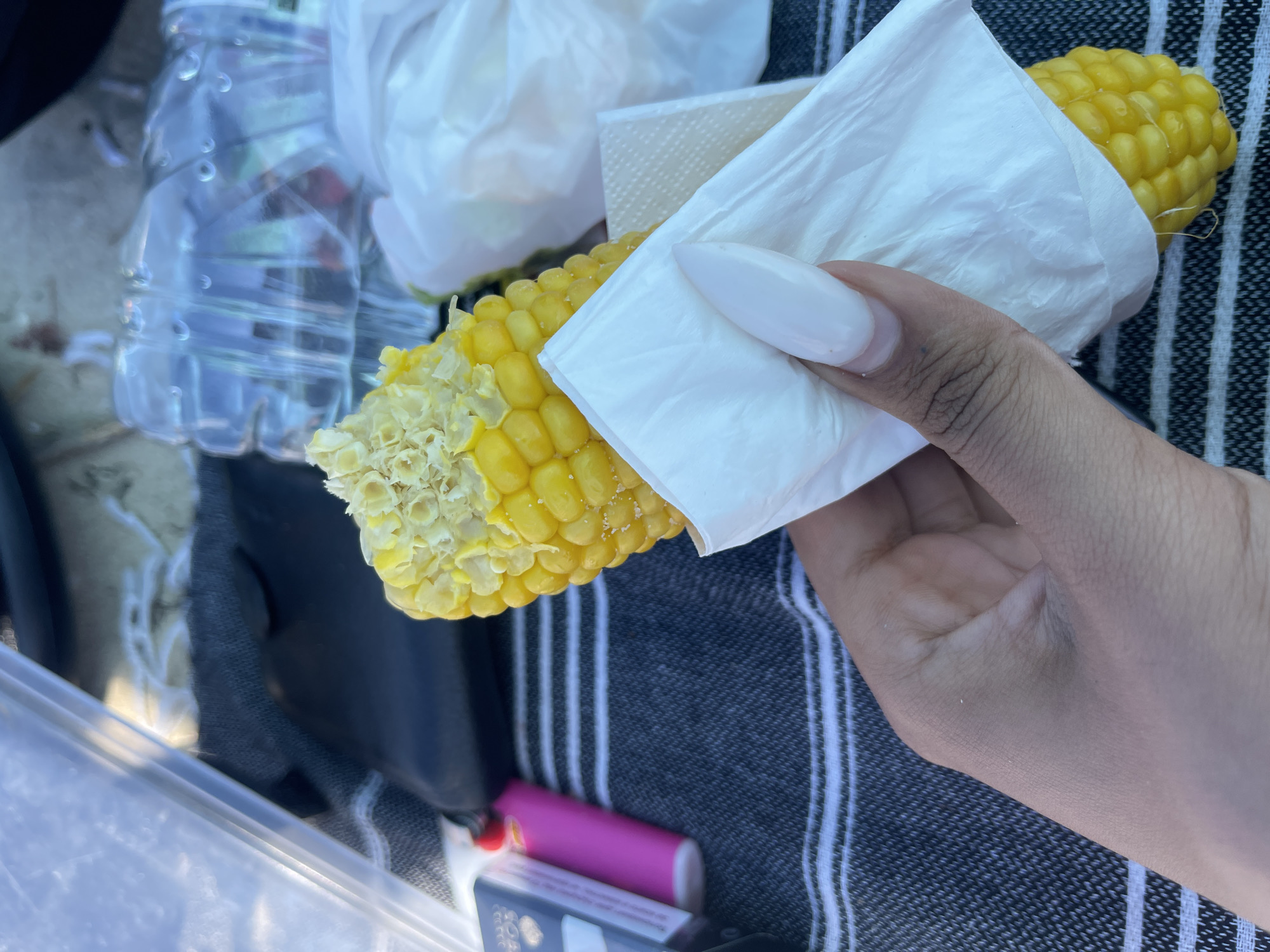 A person's hand with long, almond-shaped white nails holds a partially eaten ear of corn wrapped in a white paper napkin. The corn is bright yellow, with some kernels missing, revealing the cob. The hand is positioned in the lower right quadrant of the image, with the corn extending upwards. The background is blurred, but it appears to be an outdoor setting with a striped blanket, a clear plastic container, a pink lighter, a pack of cigarettes, and bottles of water. The image was taken in Kranevo, Bulgaria.