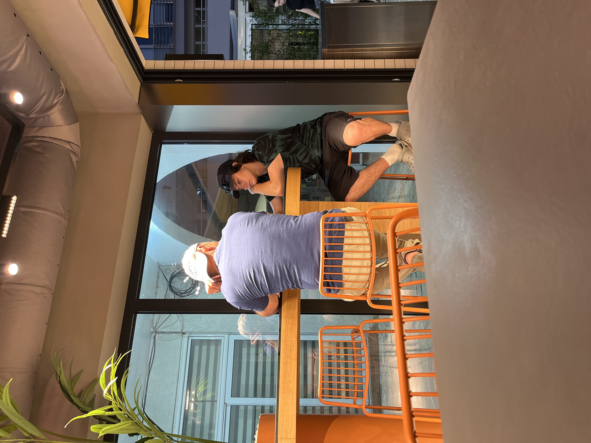 In Budva, Montenegro, two individuals are seated at a long wooden table, positioned by a large window. The person on the left, viewed from behind, wears a blue t-shirt and a white baseball cap. They are leaning on their arms on the table. The person on the right, facing left and leaning their head on their hand, wears a dark green and black patterned t-shirt, black shorts, and grey crocs with white socks. They have a black baseball cap on. In the foreground, a light brown tabletop is visible, partially obscuring the view of the people. Behind the individuals, bright orange chairs are arranged along the table. The environment appears to be a restaurant or cafe with indoor lighting, and through the window, an outdoor scene with a building facade and greenery is visible.