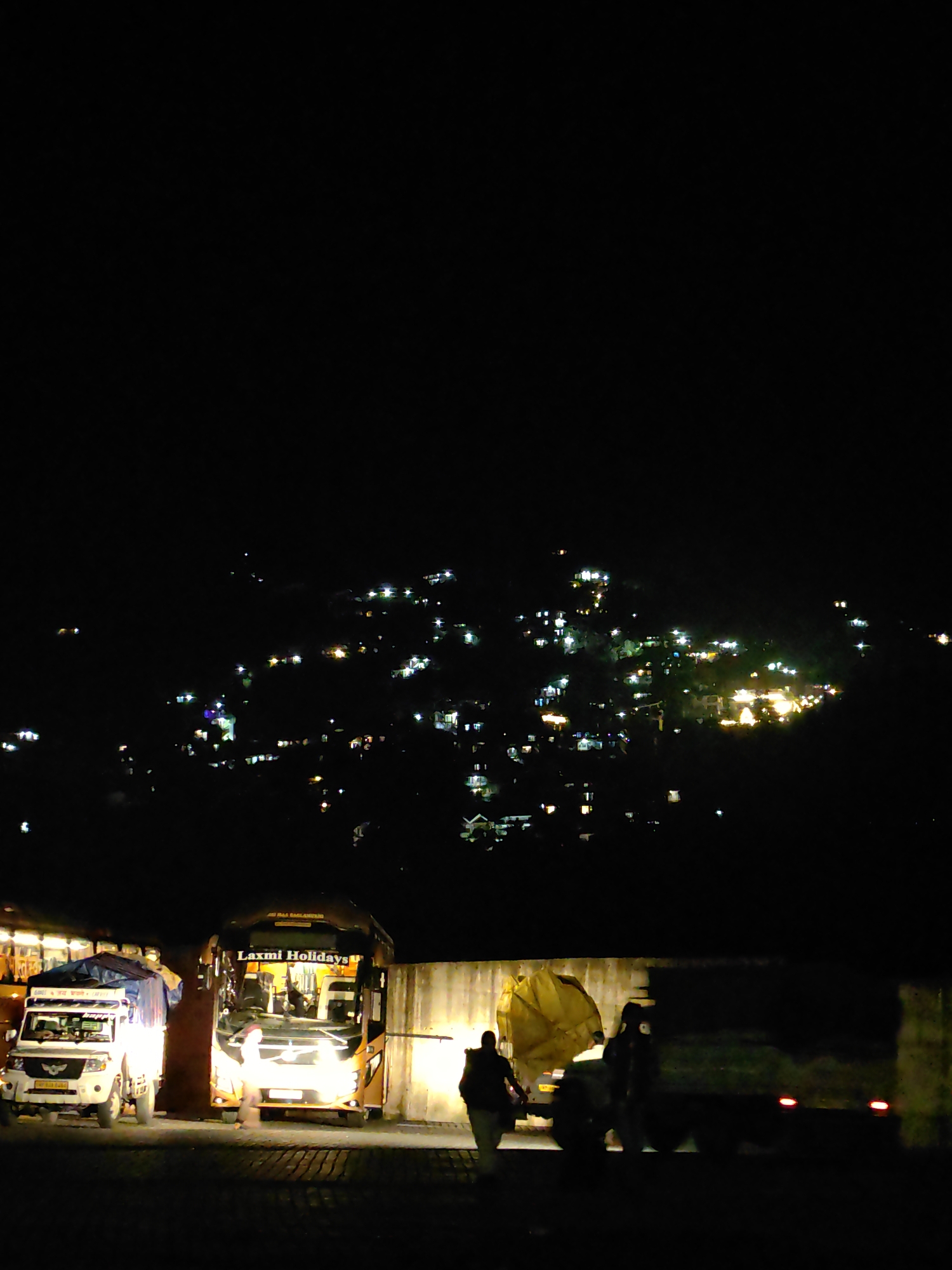 An outdoor nighttime scene depicts the lights of a town or city twinkling on a hillside in the background. In the foreground, two vehicles are illuminated by their headlights and nearby lights. On the left is a white truck with a covered cargo bed. To its right is a large orange bus with "Laxmi Holidays" written on its side. The bus has its headlights on, illuminating the area. A person is walking towards the bus from the left. Another person is standing near the right side of the bus. The ground in front of the vehicles appears to be a paved surface. The overall atmosphere is dark, with the lighting on the vehicles and distant town creating points of visibility.