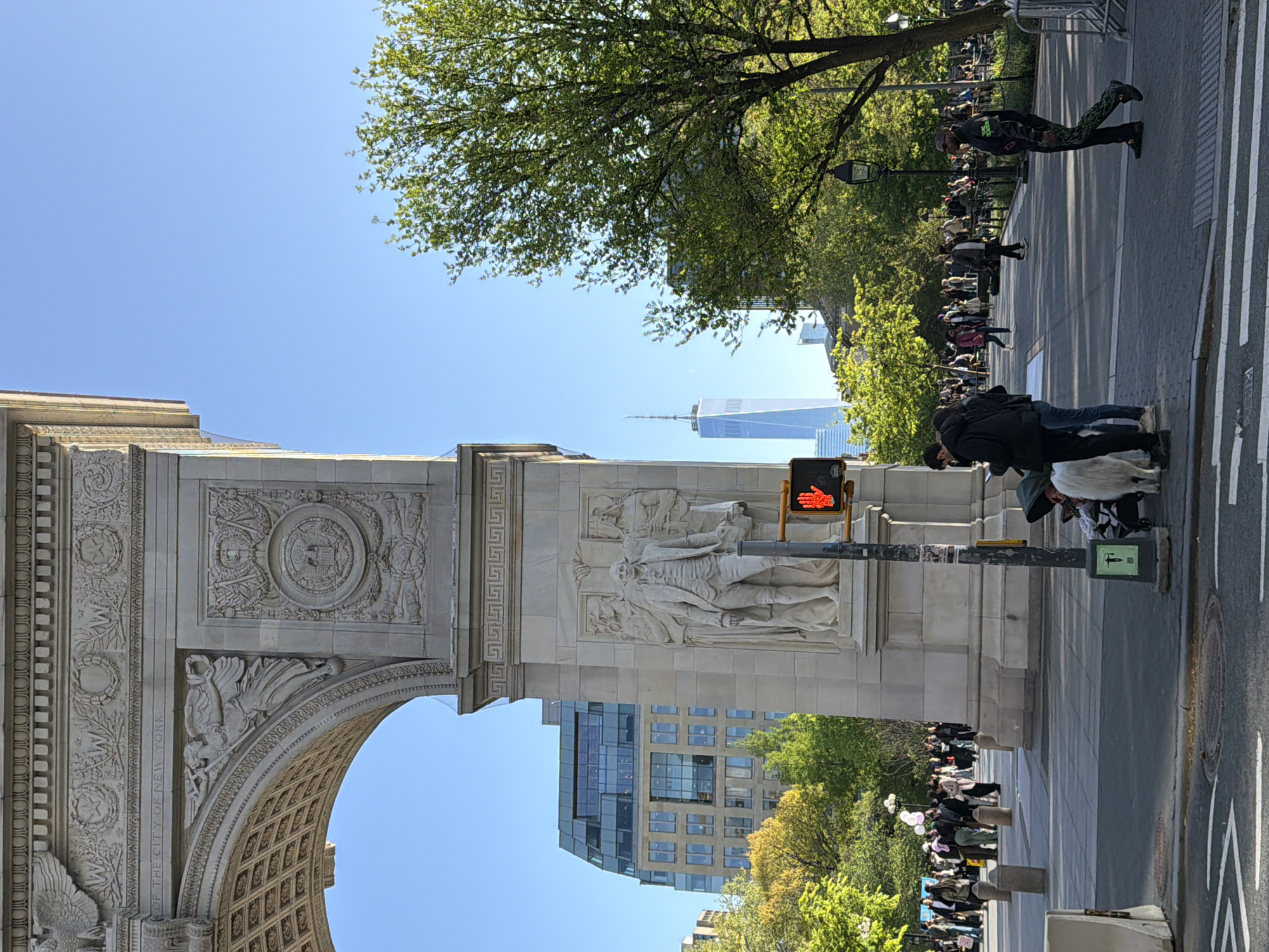The image shows the Washington Square Arch in New York City, viewed from the ground level. The arch, made of stone, features intricate carvings, including a statue of George Washington. The One World Trade Center building is visible in the background. A crowd of people is visible on the sidewalk, with some crossing the street. A pedestrian crossing signal displays a red hand indicating 'Do Not Walk'.