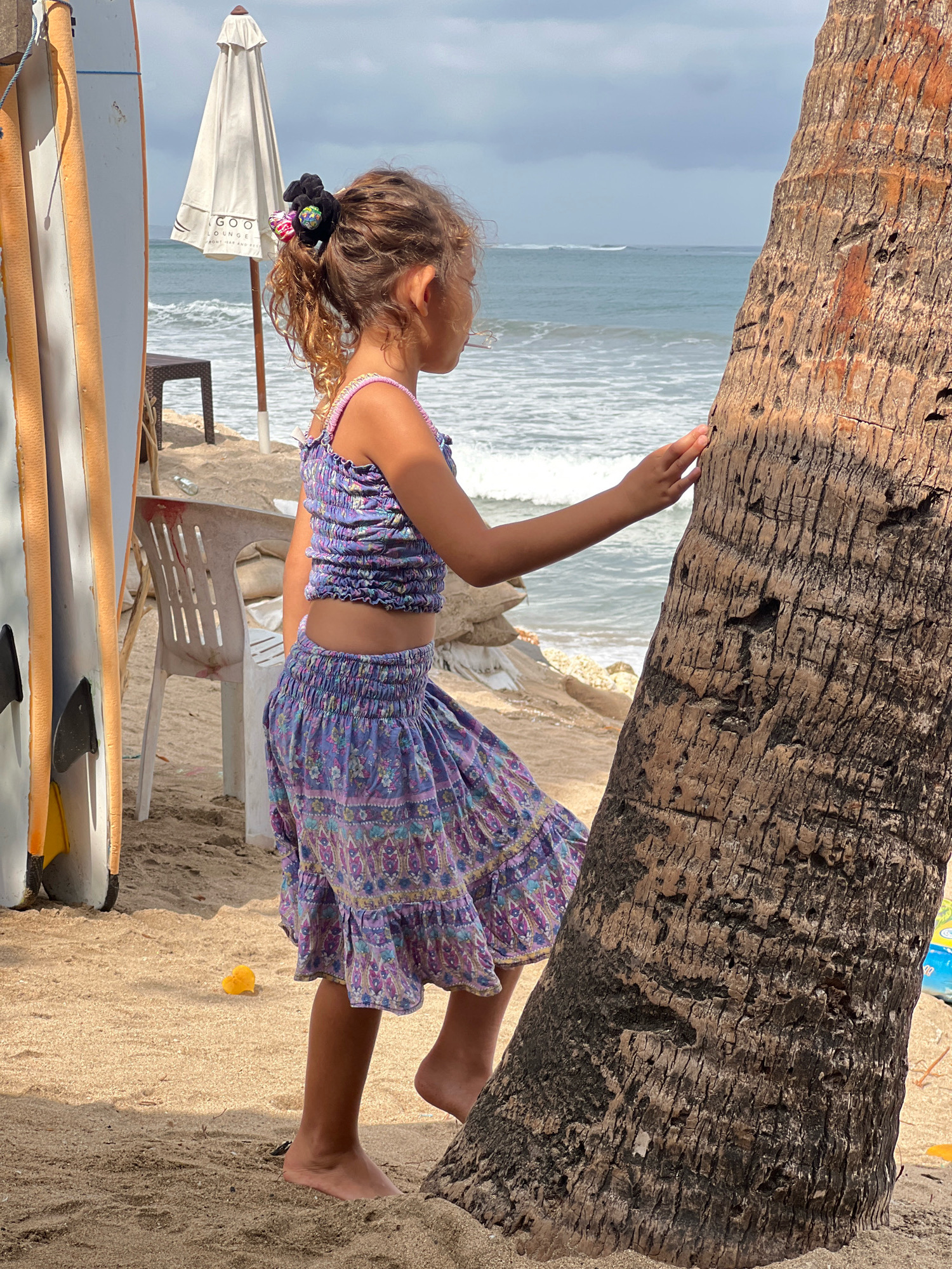 A young girl with curly brown hair pulled back with a black scrunchie with colorful accents stands on a sandy beach in Kuta, Indonesia. She is wearing a purple floral print two-piece outfit consisting of a cropped top and a ruffled skirt. The girl is facing away from the camera, looking out at the ocean, and has her right hand resting on a large palm tree trunk. The ocean is visible in the background with small waves. To the left of the girl, two surfboards stand upright. A white umbrella and a white plastic chair are also present on the beach. A small yellow object, possibly a toy, lies in the sand near the girl's feet. The lighting suggests it is daytime, with some clouds in the sky.