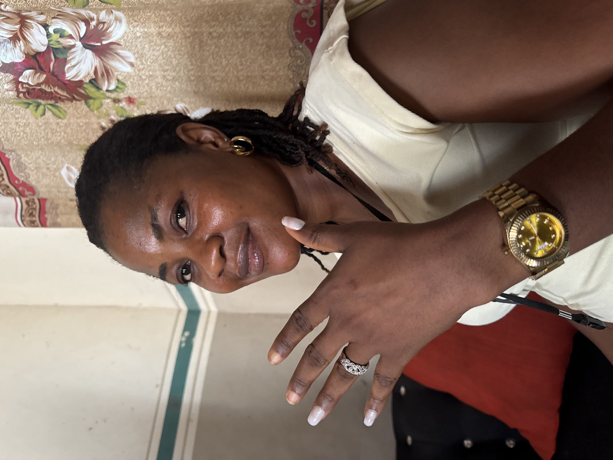 A woman with dark skin and braided hair is wearing gold earrings, a gold watch, and a diamond ring. She is wearing a light-colored shirt. She is in front of a floral patterned surface. The setting cannot be confirmed as Badagry, Nigeria without additional information.