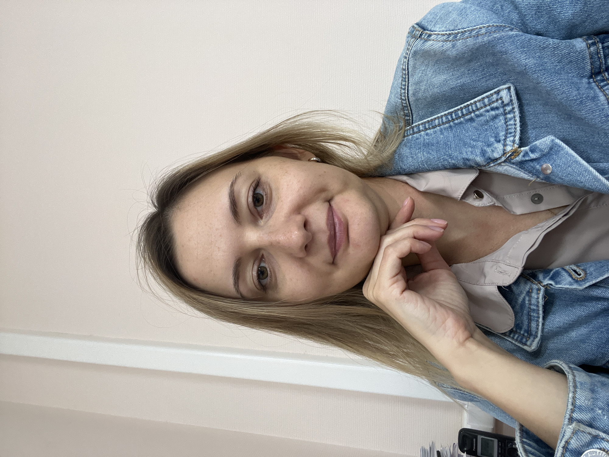 A selfie of a Caucasian woman with blonde hair and light skin, wearing a light pink button-down shirt and a blue denim jacket, is taken indoors. She is resting her chin on her hand, and her gaze is directed at the camera. A light beige wall serves as the backdrop. To the left of the frame, a portion of a wall with a white vertical trim is visible, along with a black cordless phone and a stack of papers. The scene is consistent with an office environment. It is not possible to determine the specific location within the city of Volgograd, Russia, from the provided image.
